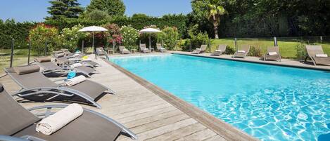 Zona de piscina con sillones de descanso y sombrillas, rodeada de follaje.