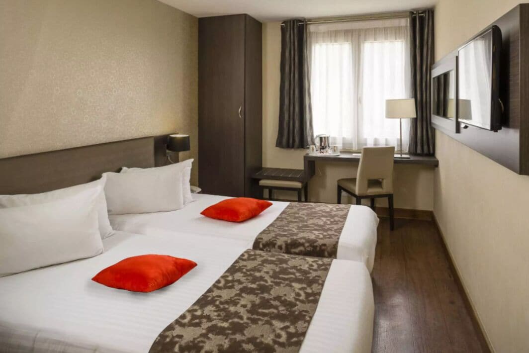 A hotel room with two beds, a desk, a chair, a TV, and a window with curtains.