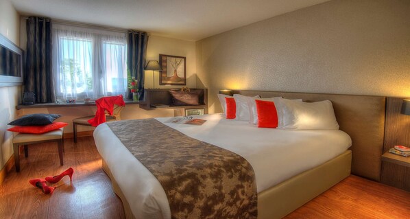 Quarto de hotel com uma cama grande, uma escrivaninha, uma televisão e uma janela com cortinas.