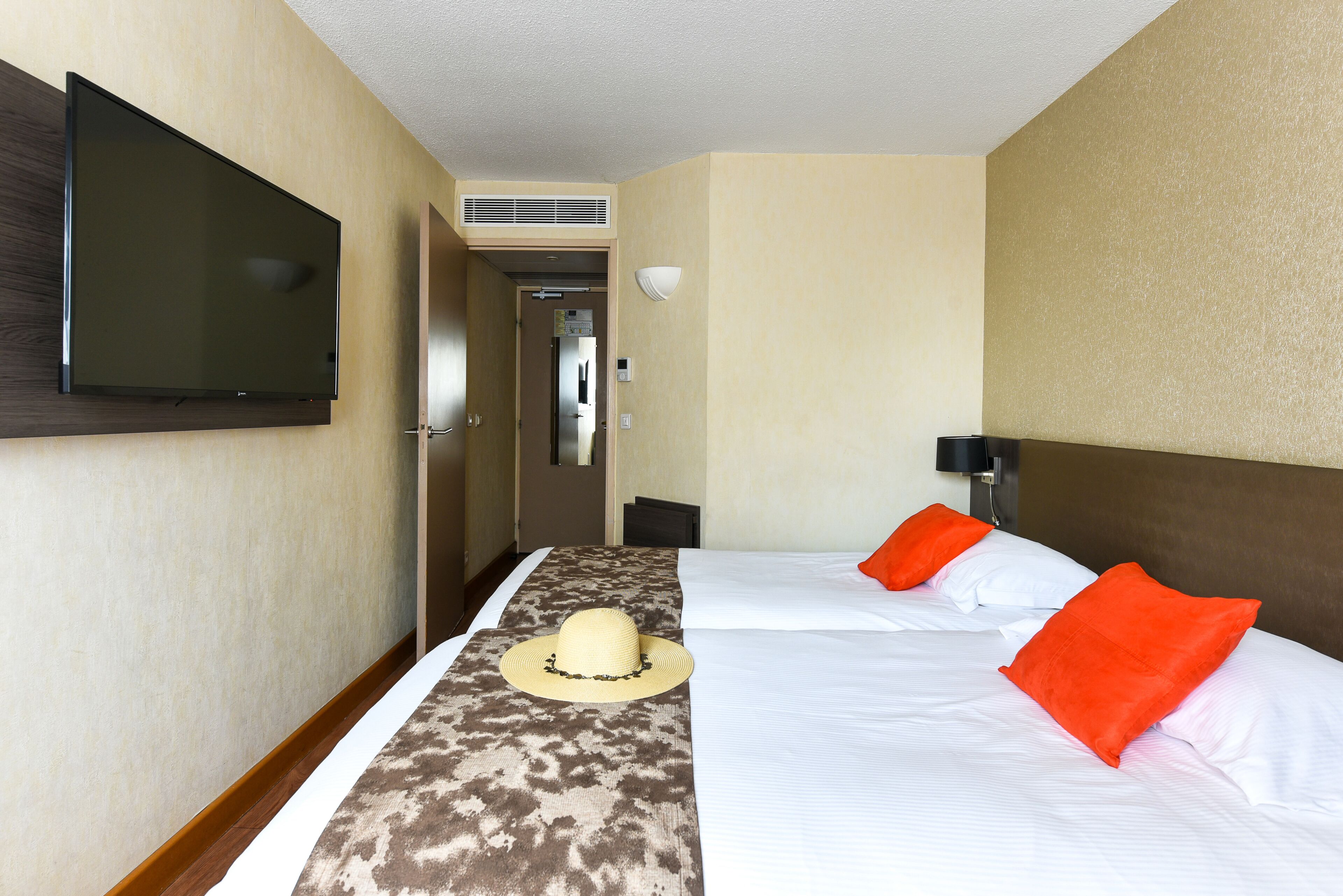 Een hotelkamer met twee bedden, een aan de muur bevestigde televisie en een deur naar een gang.