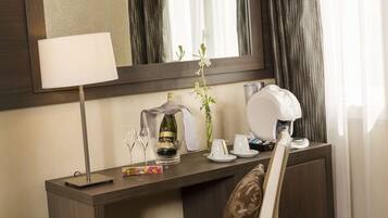 A hotel room with a wooden desk, a mirror, a lamp, a bottle of champagne, a vase with a flower, and a coffee maker.