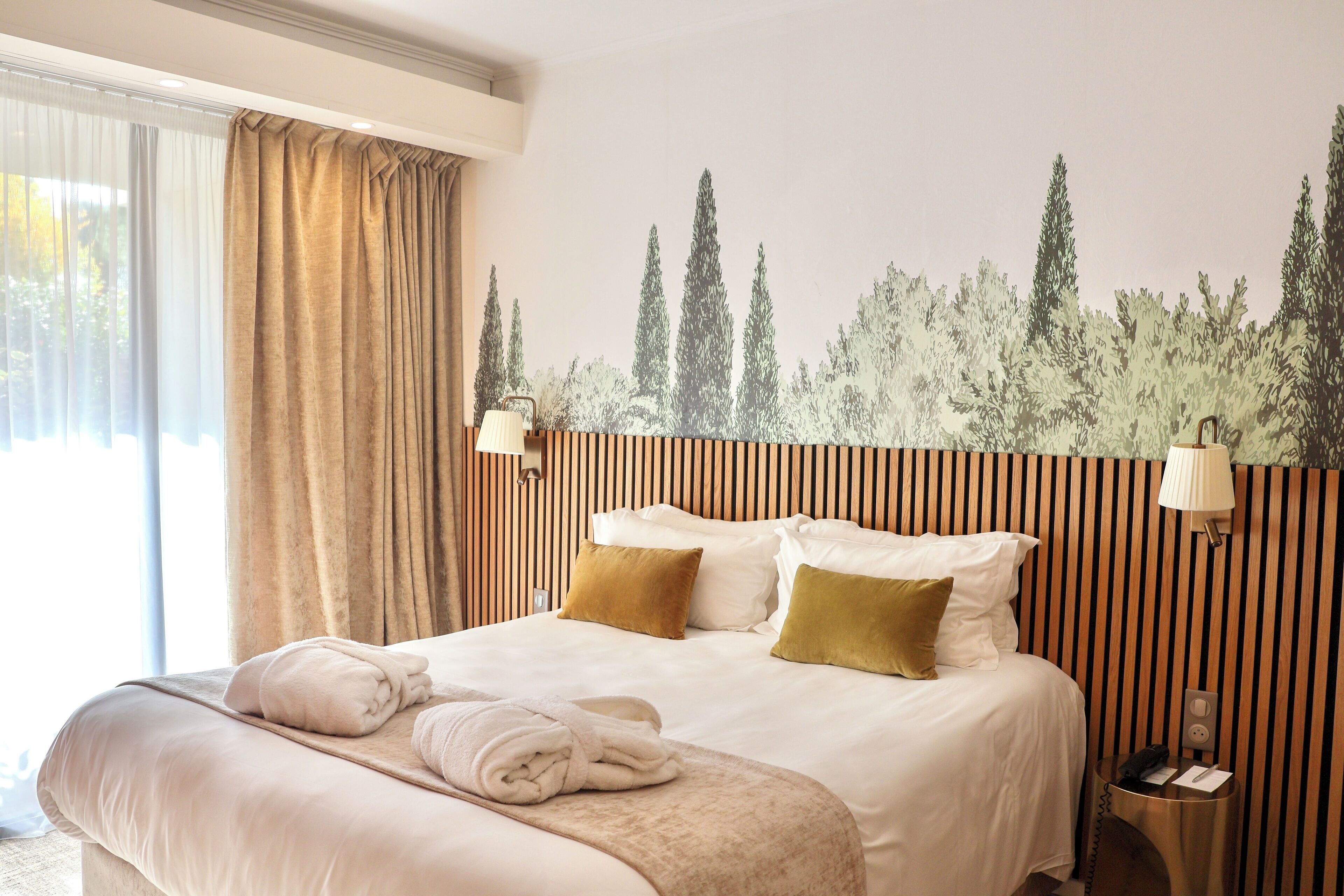 A hotel room with a large bed, two pillows, a bedside table with a lamp, and a wall mural of trees.