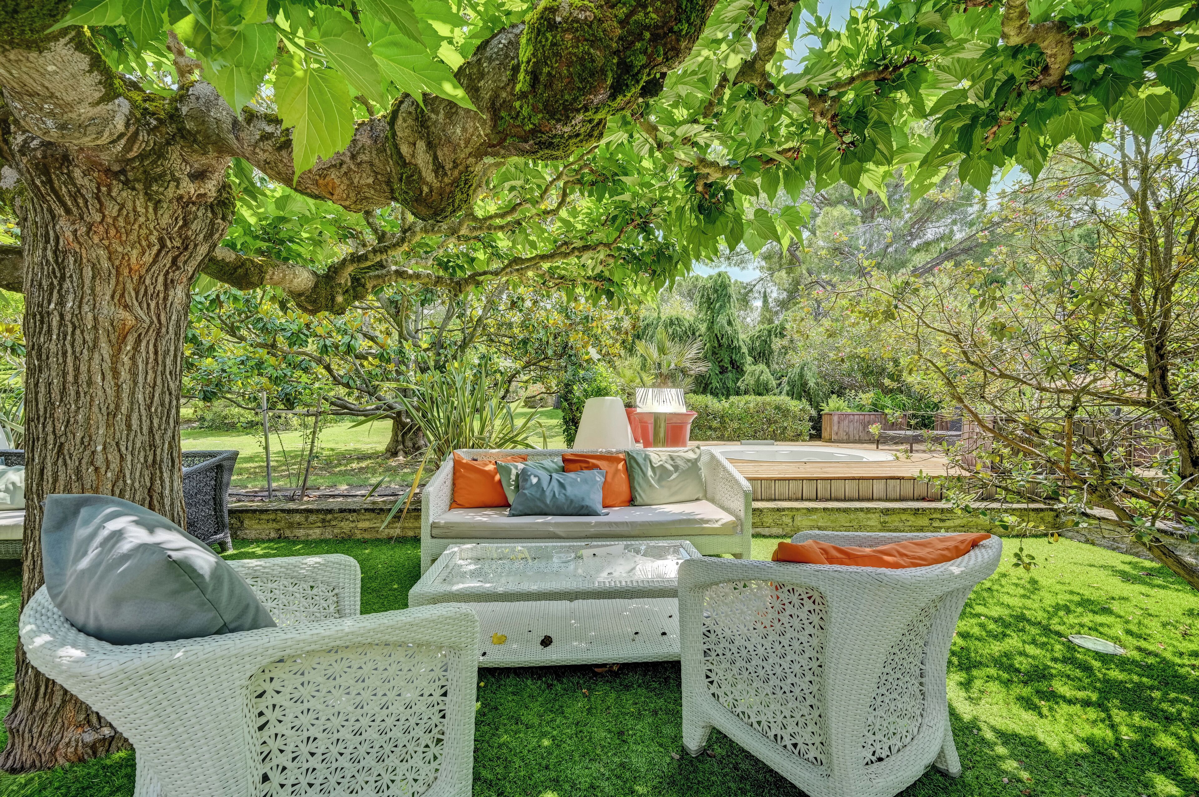 Un espace patio aménagé avec des meubles en osier, un canapé blanc agrémenté de coussins orange et un grand arbre qui offre de l’ombre.