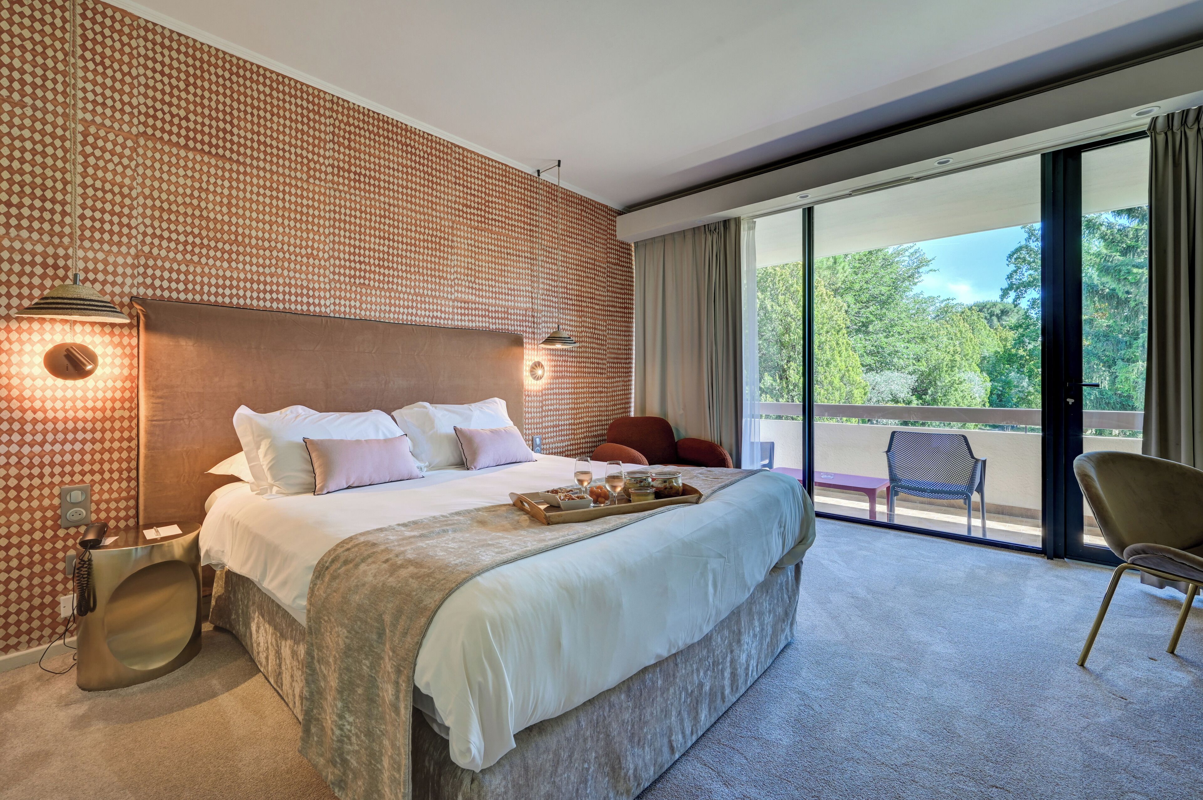 A modern hotel room with a large bed, a balcony with a view of greenery, and a small seating area.