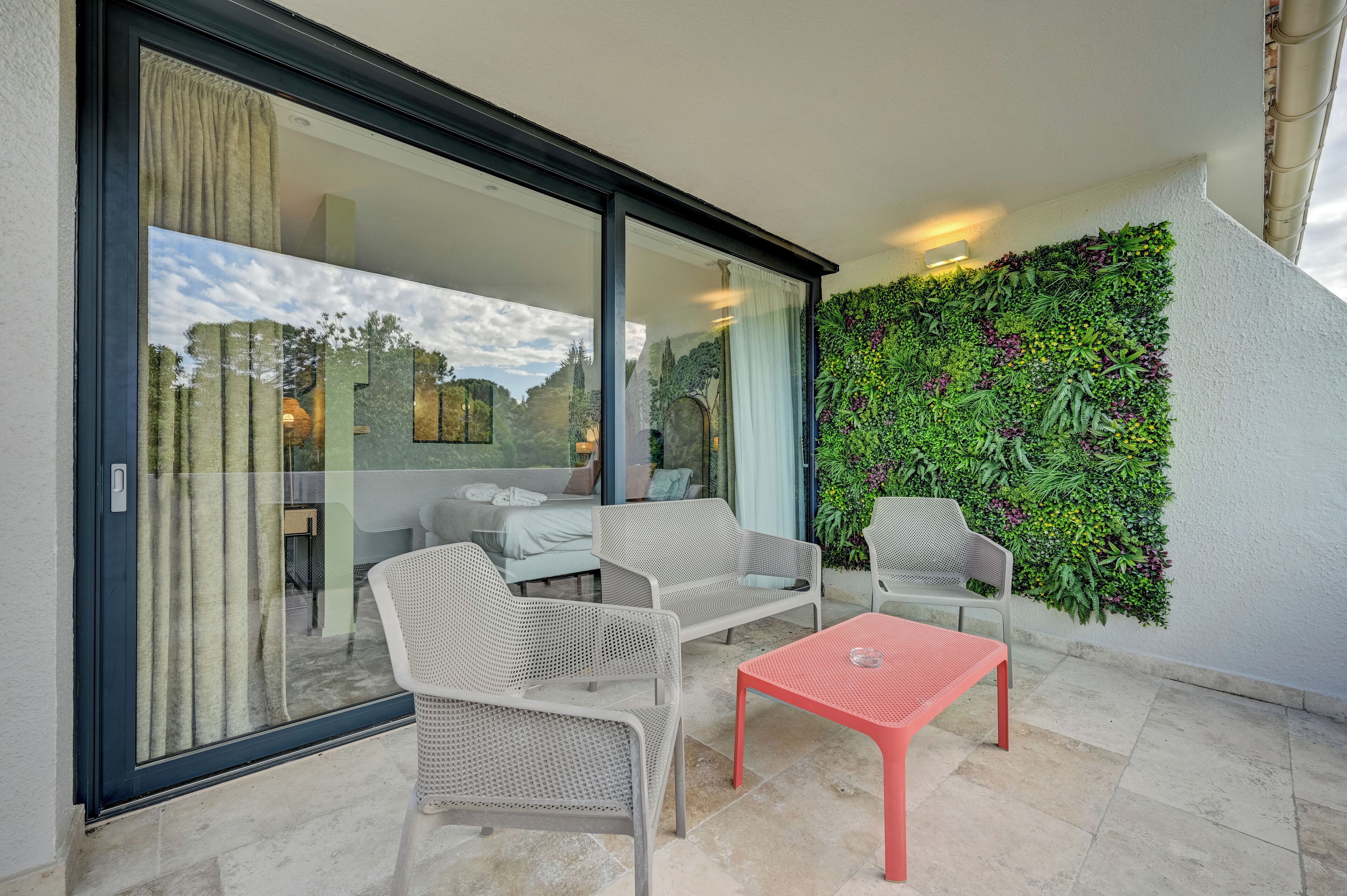 A balcony with wicker furniture, a red table, and a green vertical garden.