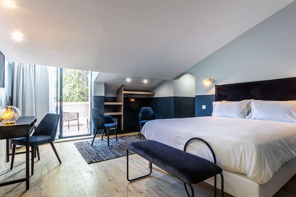 A modern hotel room with a large bed, a desk with a chair, a bench, and a view of the outdoors through a sliding door.