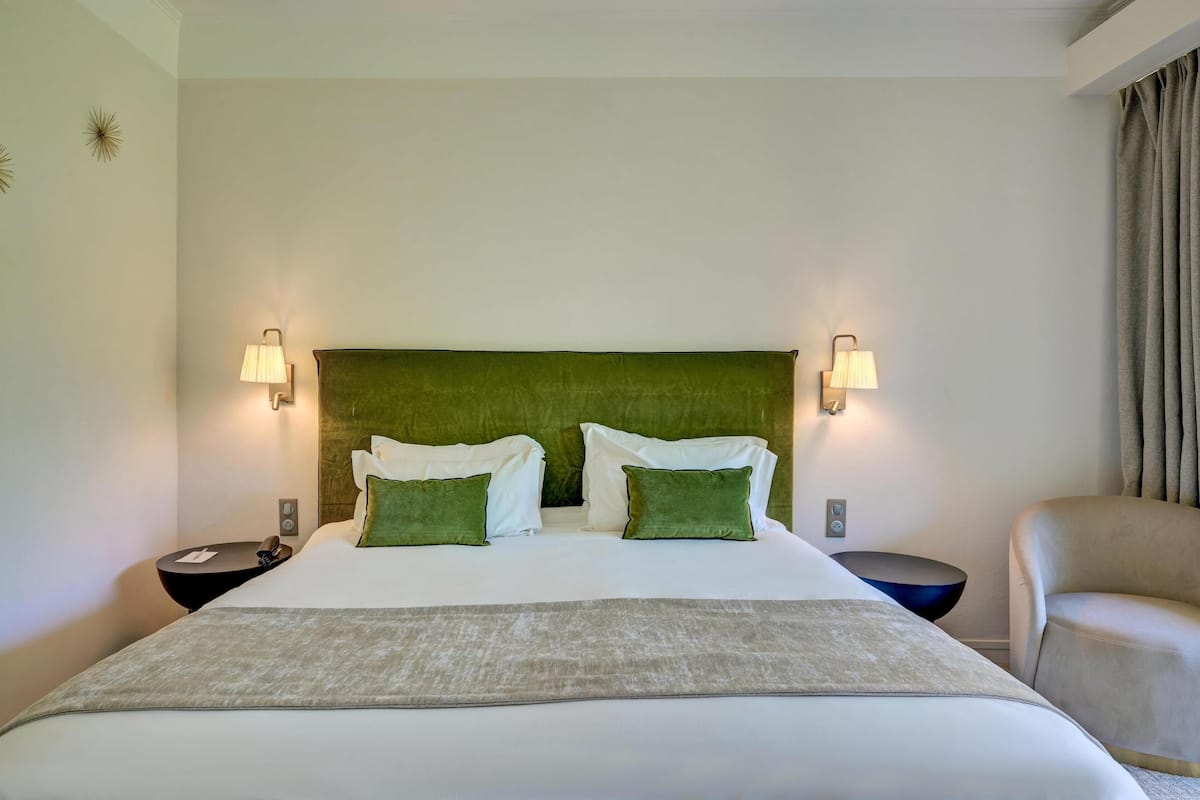 A hotel room with a large bed, two bedside lamps, a green upholstered headboard, and a beige armchair.