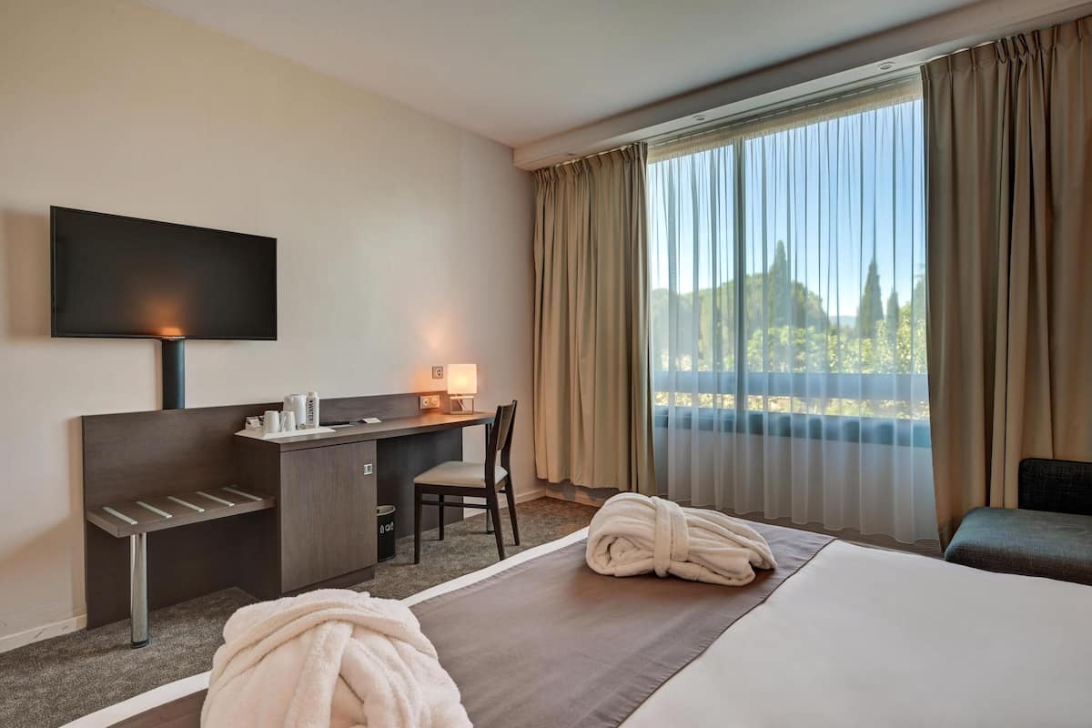 A hotel room with a bed, a desk with a chair, a TV, and a window with curtains.