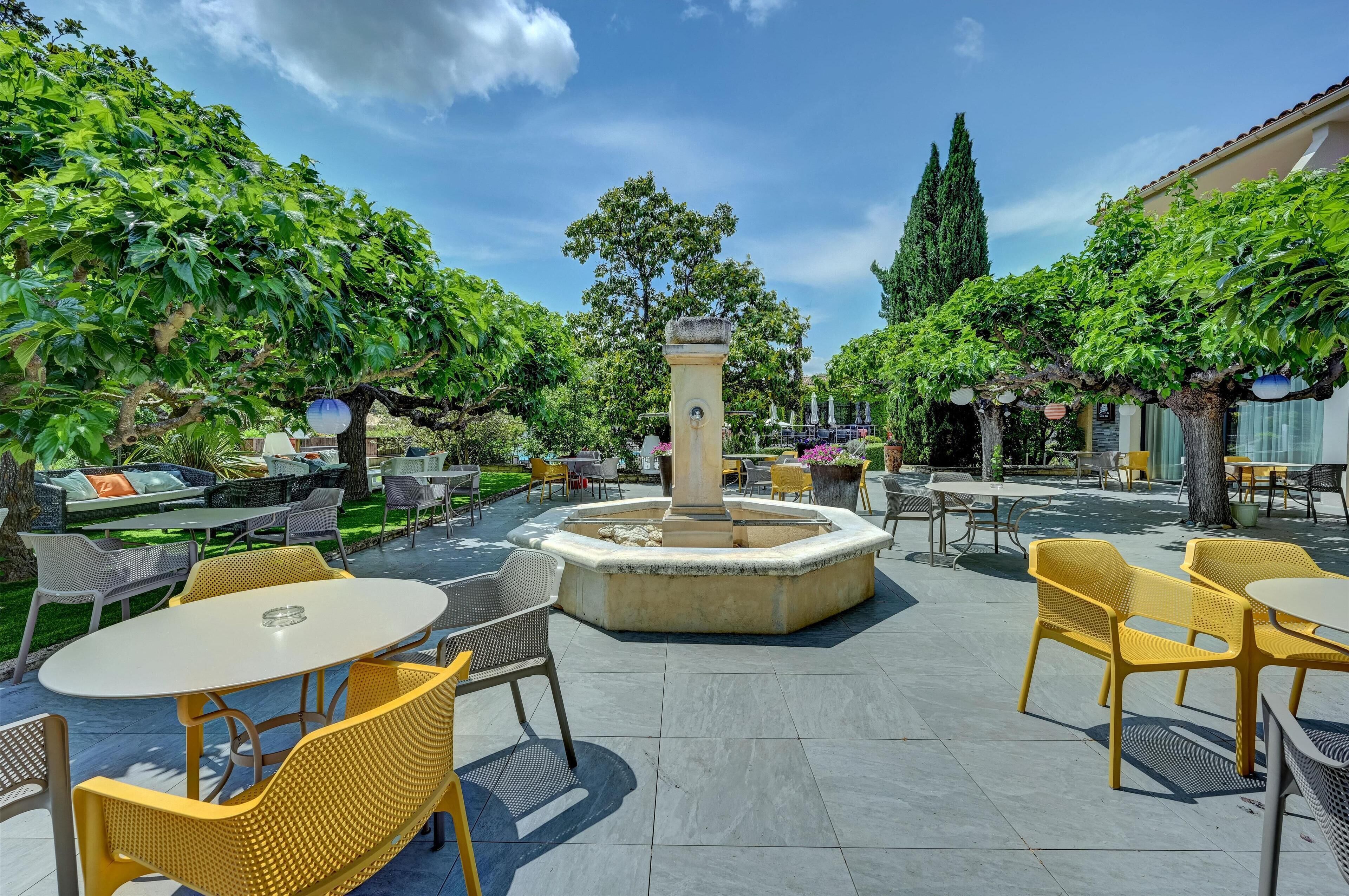 An outdoor patio area with round tables and yellow chairs, surrounded by greenery and a central fountain.