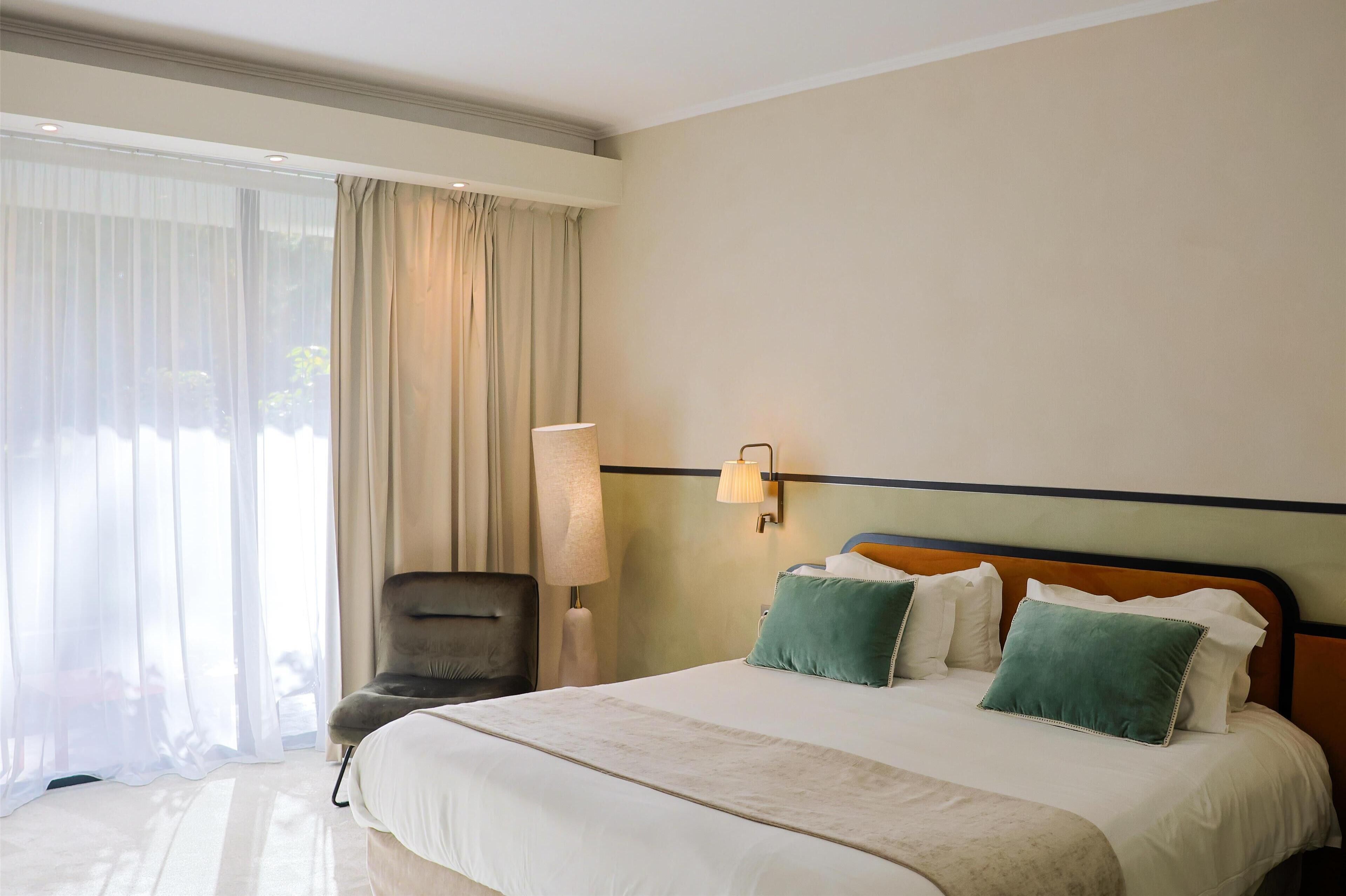 A hotel room with a large bed, a chair, a lamp, and a view through sheer curtains.