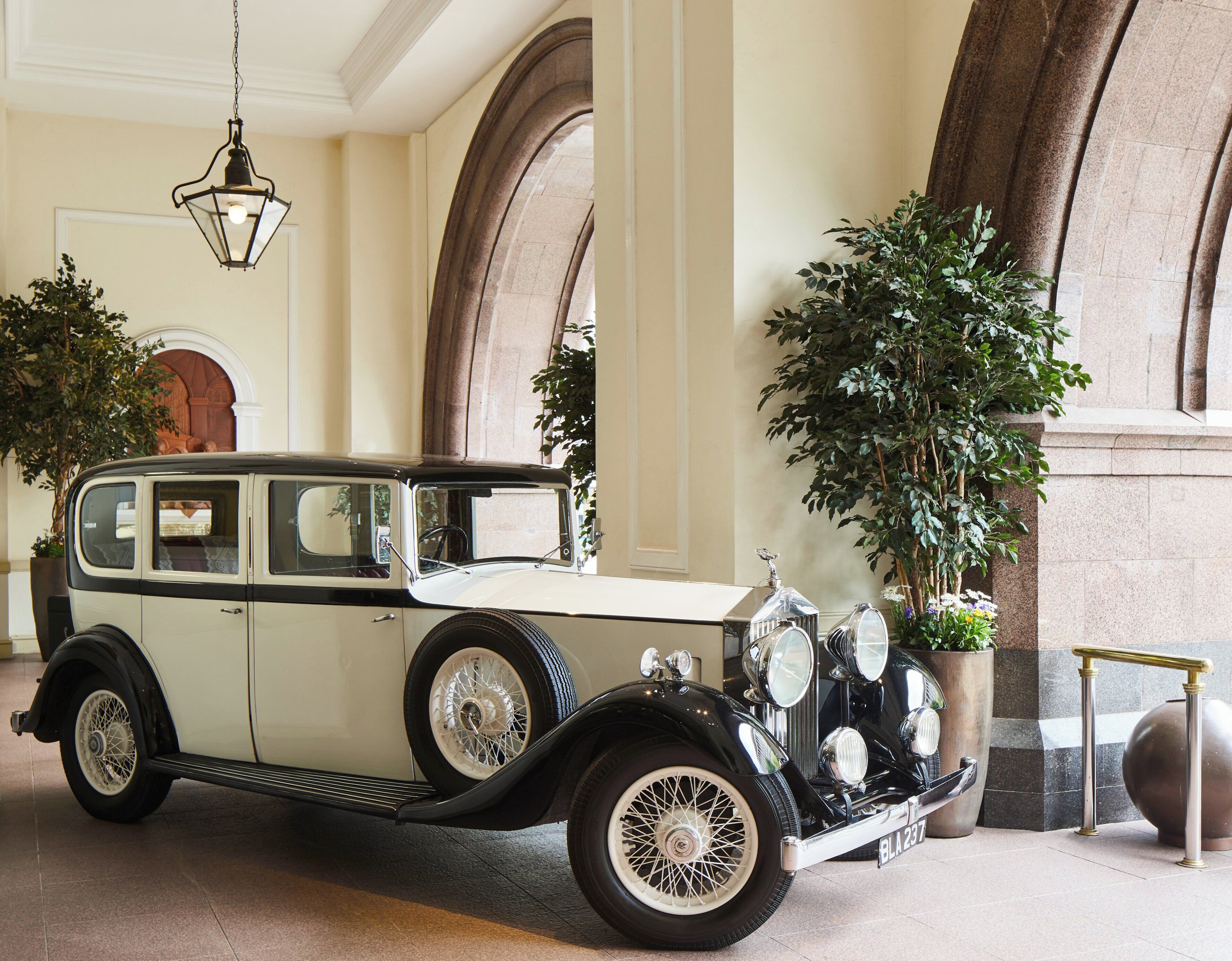 Un coche antiguo estacionado en el interior, con amplios ventanales, una araña de cristal y plantas en macetas.