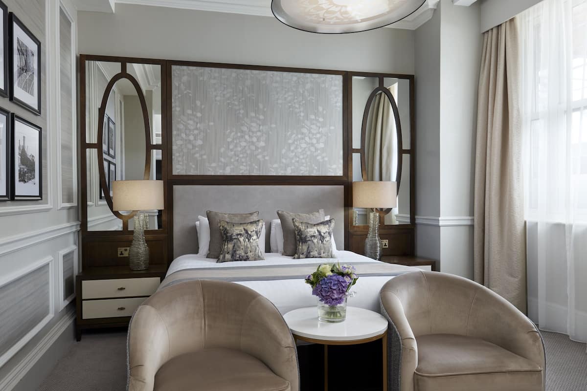 A hotel room with a large bed, two side tables with lamps, a round coffee table, two armchairs, and a decorative mirror above the bed.