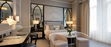 A hotel room with a bed, two armchairs, a desk with a lamp, a mirror, and a small table with a vase of flowers.