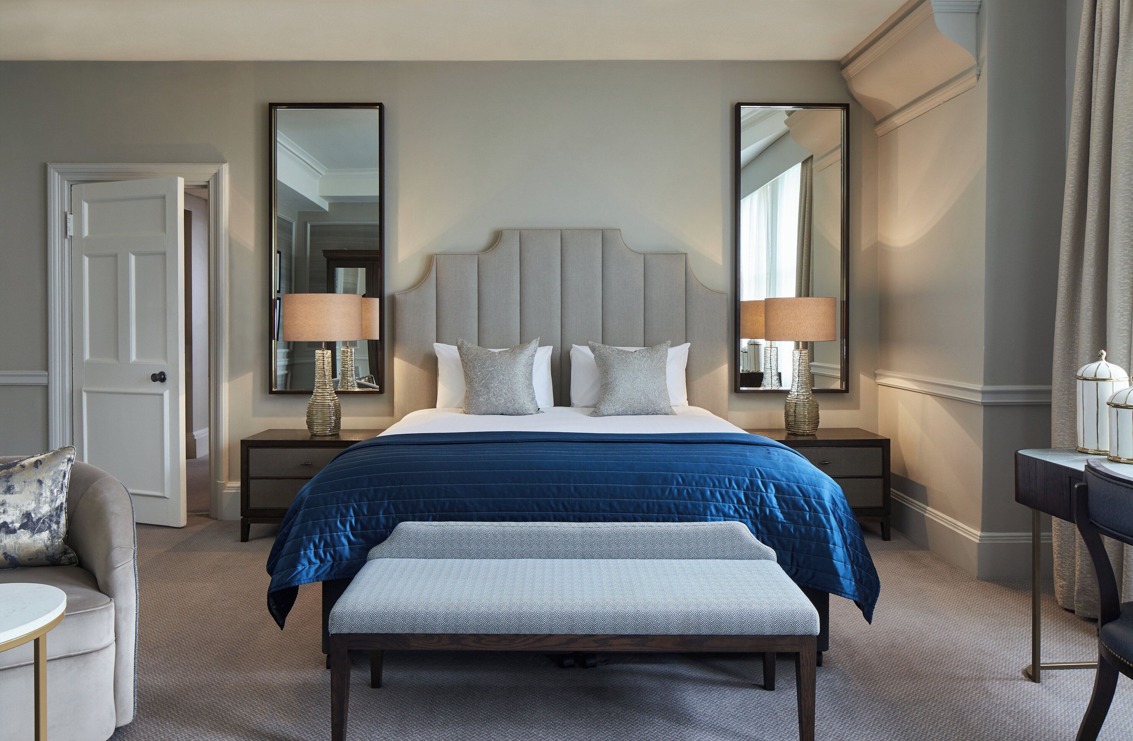A hotel room with a large bed, two bedside tables with lamps, a bench at the foot of the bed, a chair, and a desk with a lamp.