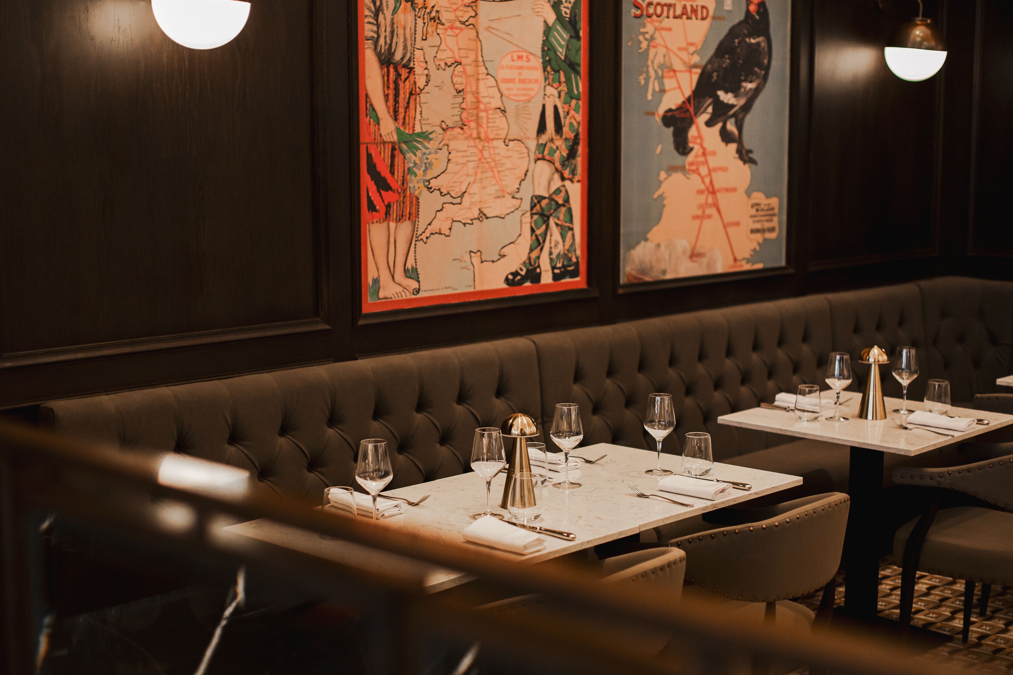 L'intérieur d'un restaurant présente un long banc rembourré, des tables dressées avec des verres à vin et des couverts, ainsi que deux appliques murales.