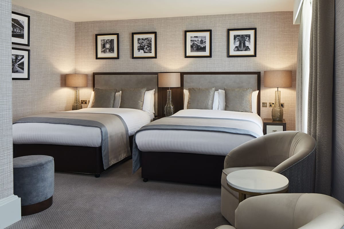A hotel room with two beds, a round table, a chair, and framed pictures on the wall.