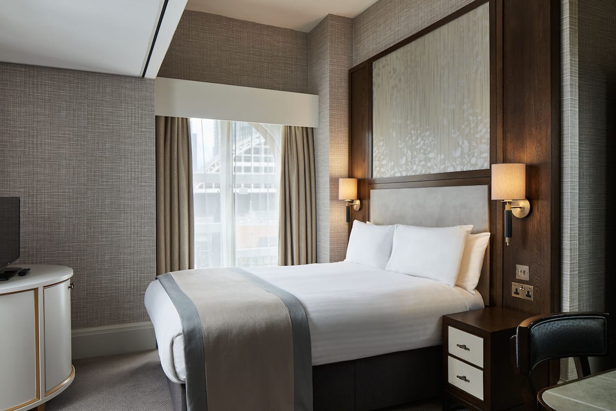 A hotel room with a large bed, a desk, and a chair. There is a window with curtains, a wall-mounted lamp, and a television on the desk.