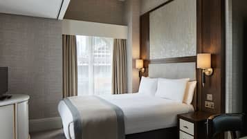 Habitación de hotel con una cama grande, un escritorio y una silla. Hay una ventana con cortinas, una lámpara de pared y un televisor sobre el escritorio.