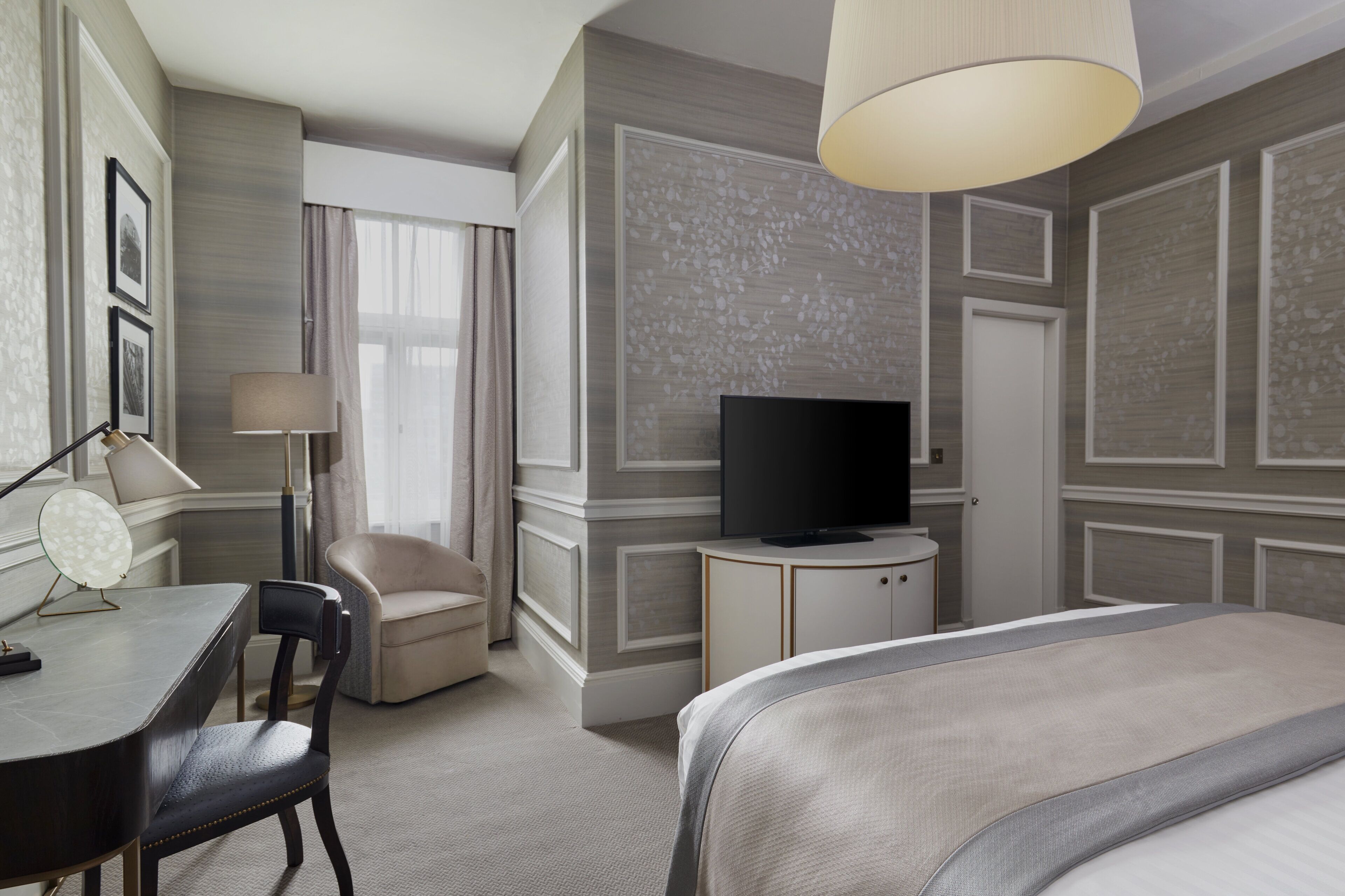 Ein Hotelzimmer mit einem Bett, einem Schreibtisch mit Lampe, einem Sessel, einem Fernseher und einem großen Fenster mit Vorhängen.