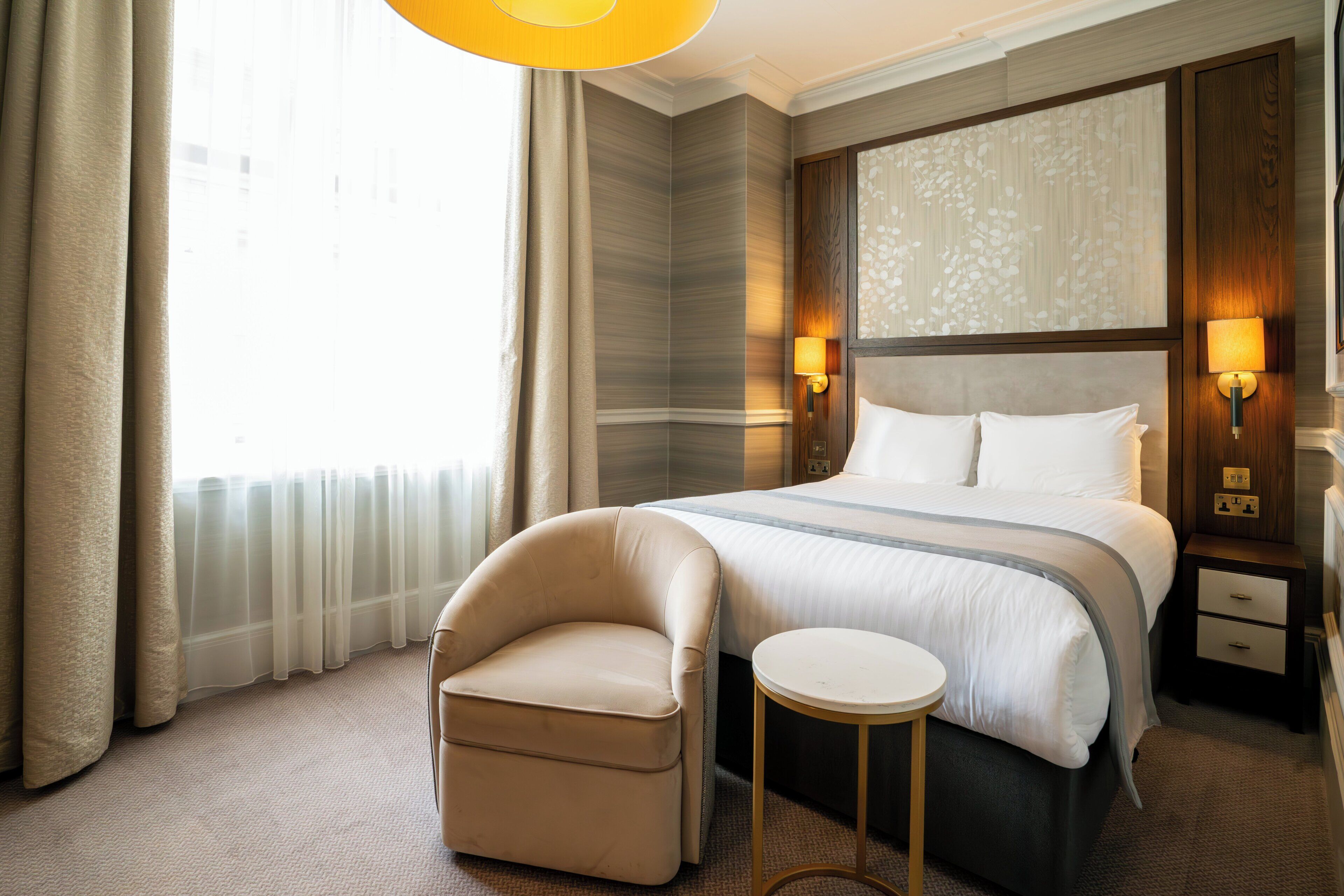 A hotel room with a large bed, a small armchair, a bedside table, and a nightstand with a lamp.