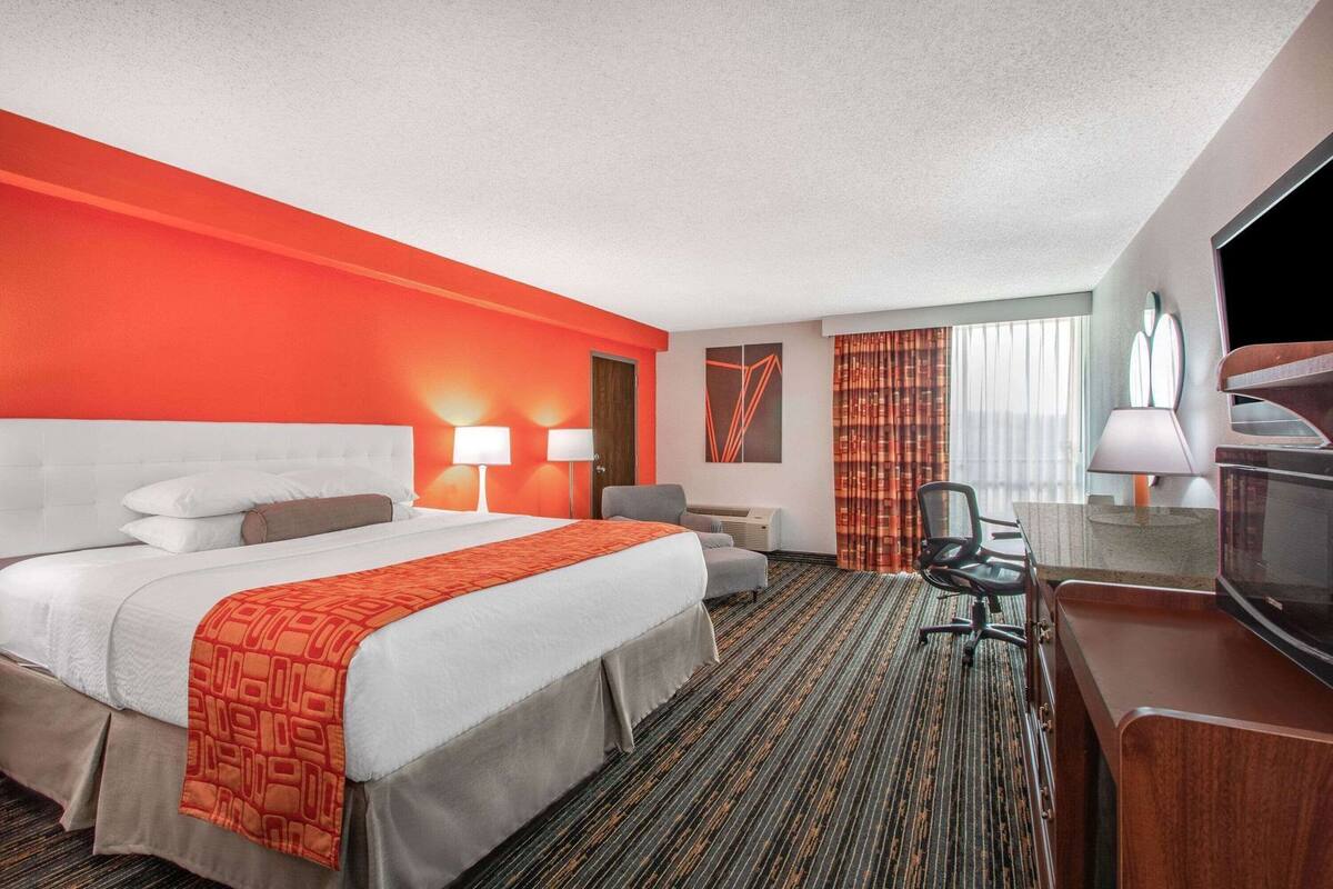 A hotel room with a large bed, two bedside tables with lamps, a desk with a chair, a television, and a window with curtains.