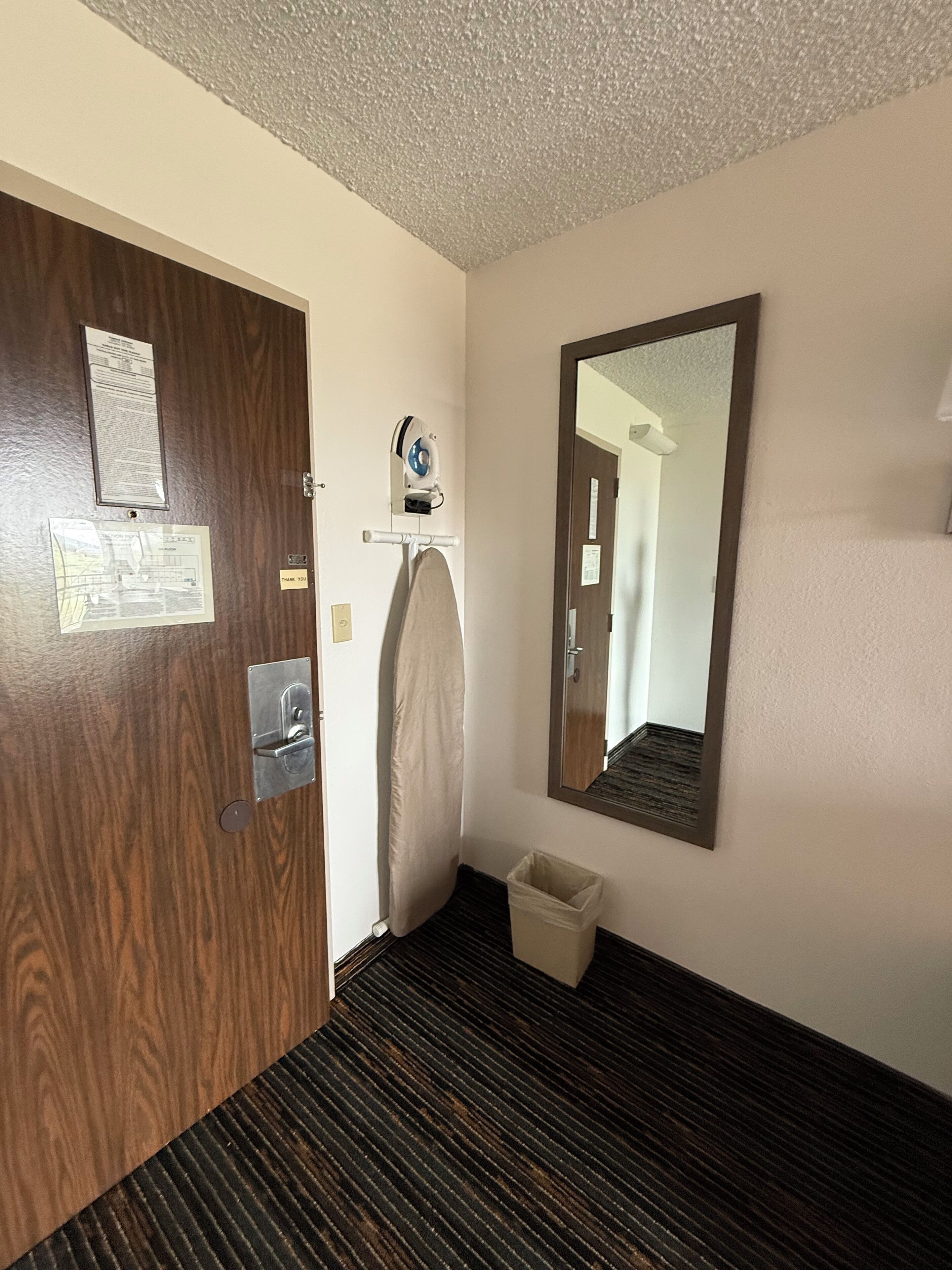 A hotel room with a wooden door, a mirror, a towel rack, and a trash bin.