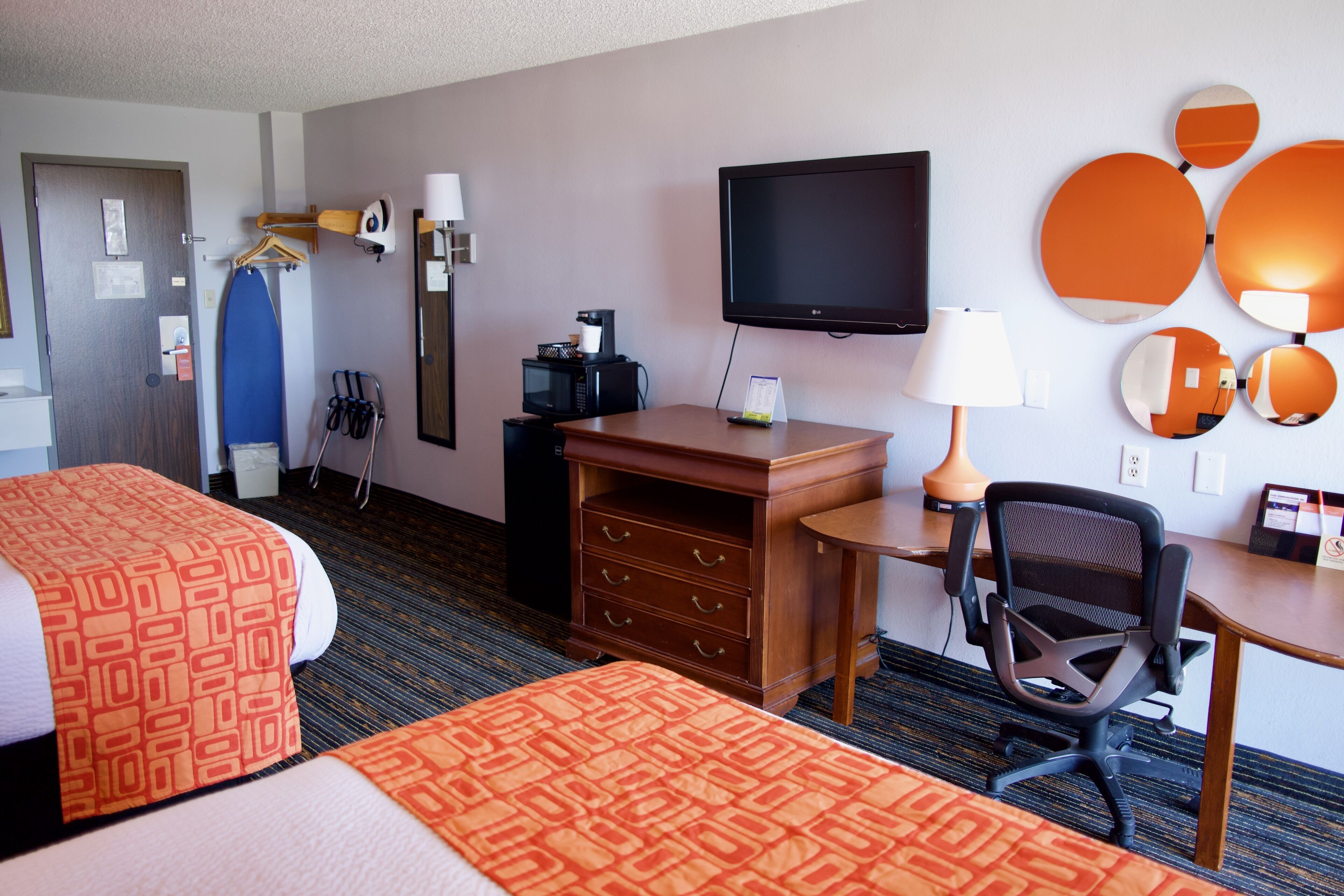 Quarto de hotel com duas camas, uma escrivaninha com cadeira, televisão, cafeteira e espelho com detalhes circulares.