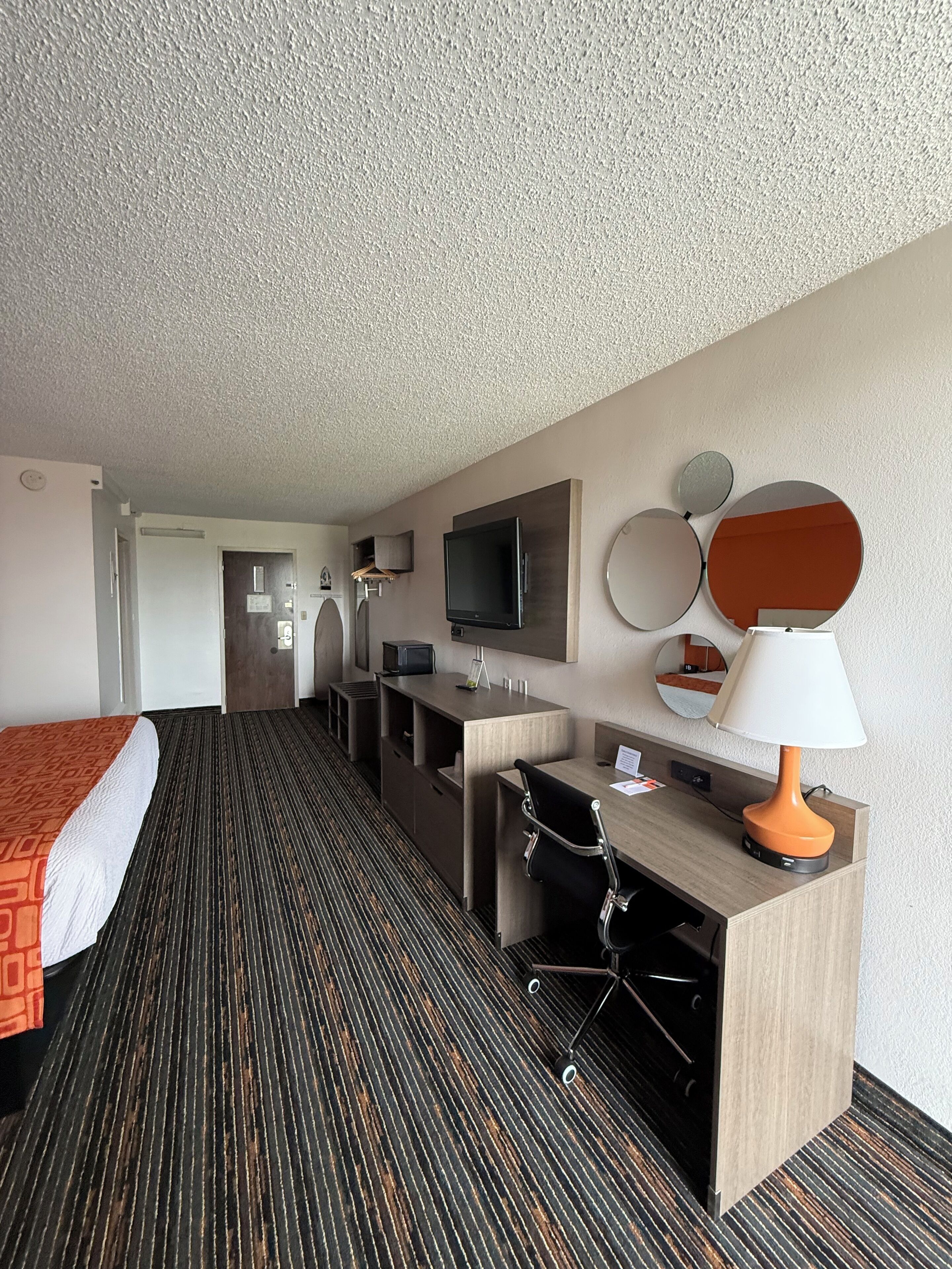 Quarto de hotel com cama, escrivaninha, cadeira, televisão e abajur.