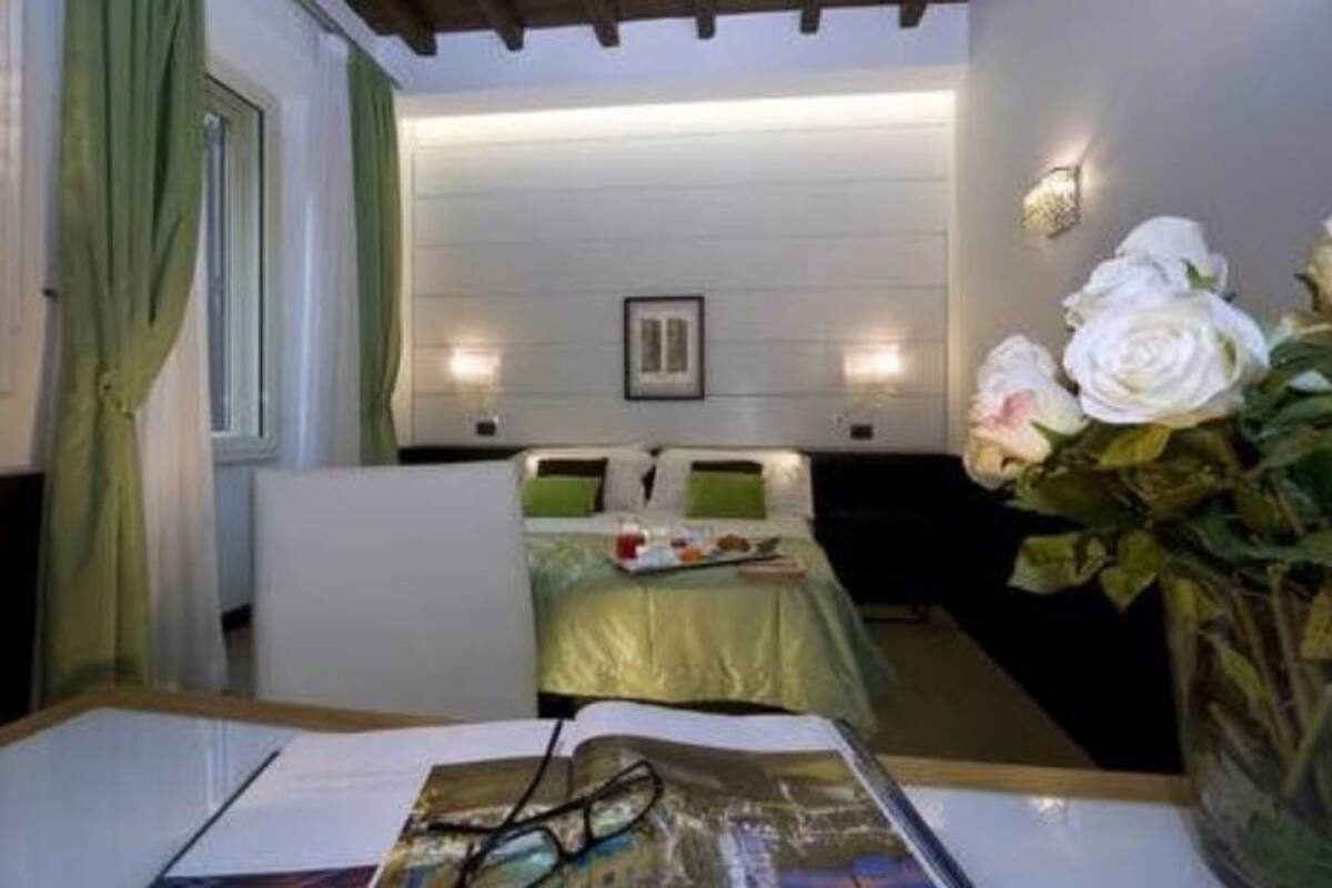 A hotel room with a bed, a bedside table with a magazine and glasses, a vase with flowers, and a wall-mounted light fixture.