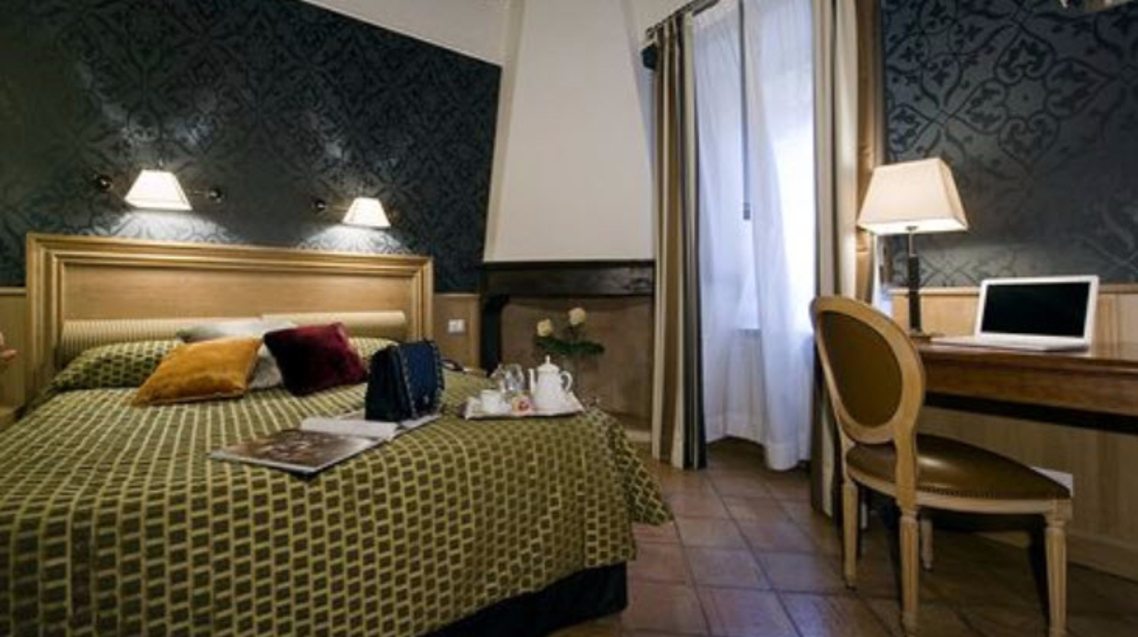 Una habitación de hotel con cama, escritorio con computadora portátil, silla y ventana con cortinas.