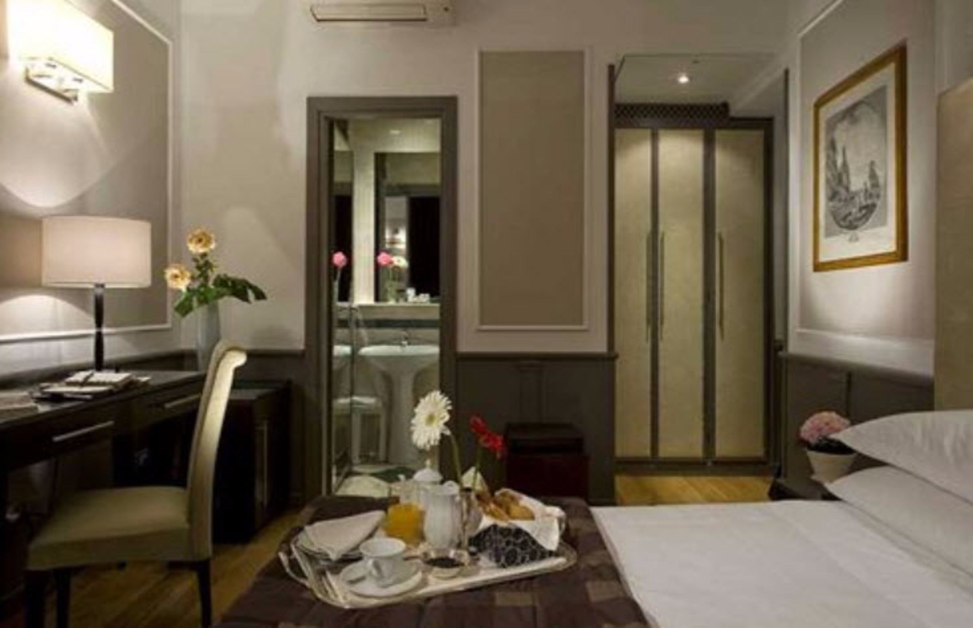 A hotel room with a bed, a desk with a lamp, a chair, a tray with breakfast items, and a framed picture on the wall.
