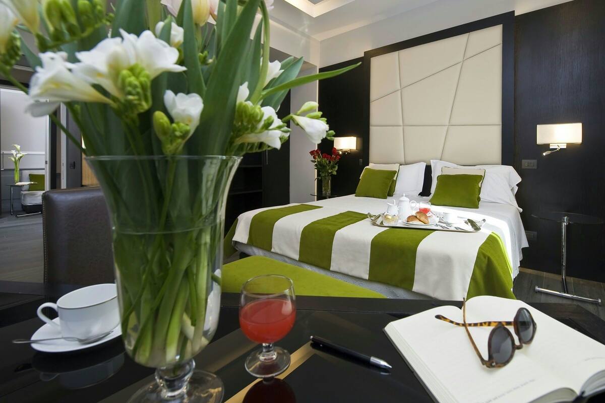 A hotel room with a bed, a dining table with breakfast items, a vase with flowers, and a view of the cityscape.