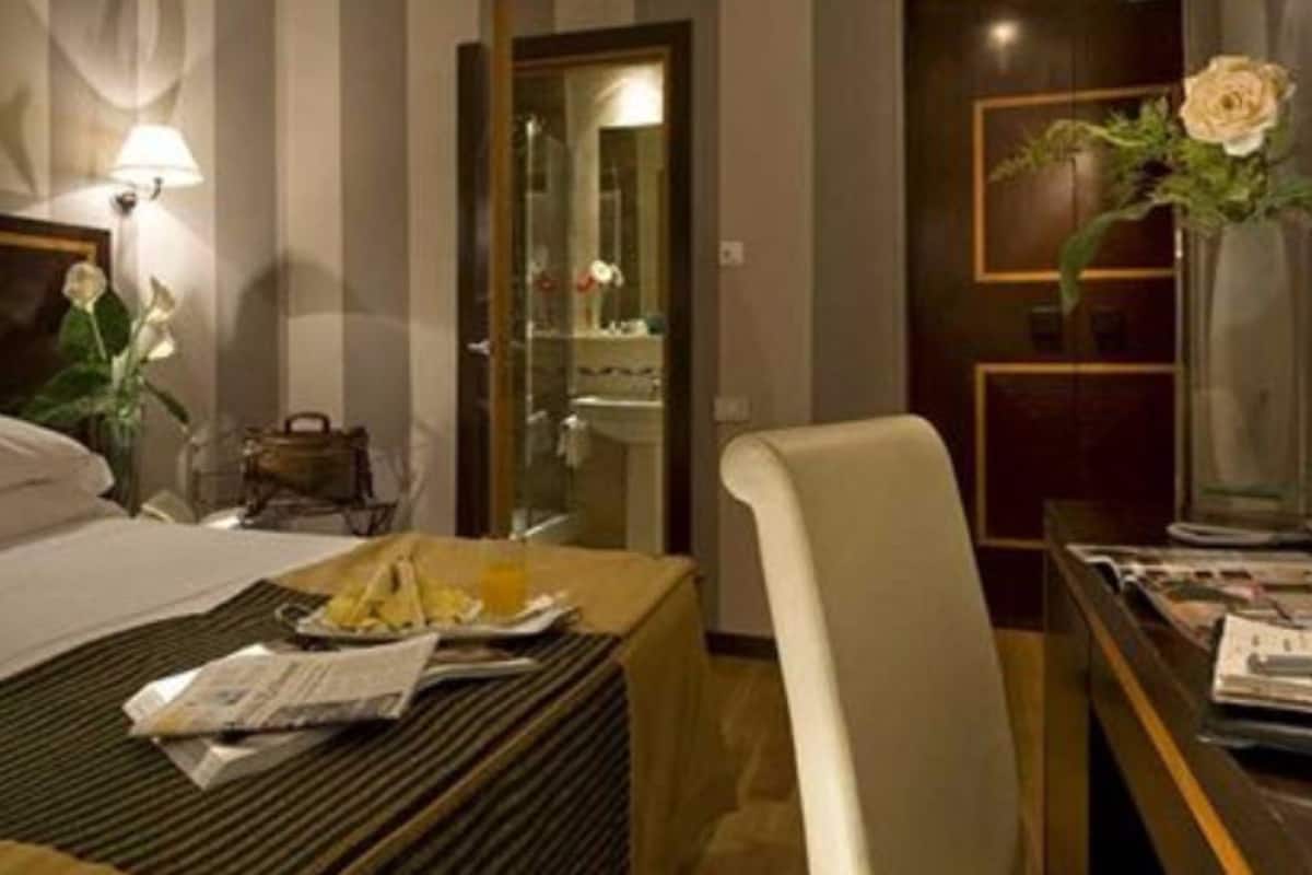 Une chambre d’hôtel comprenant un lit, une table de chevet avec une lampe, une chaise, un bureau agrémenté d’un vase de fleurs et donnant sur la salle de bain.