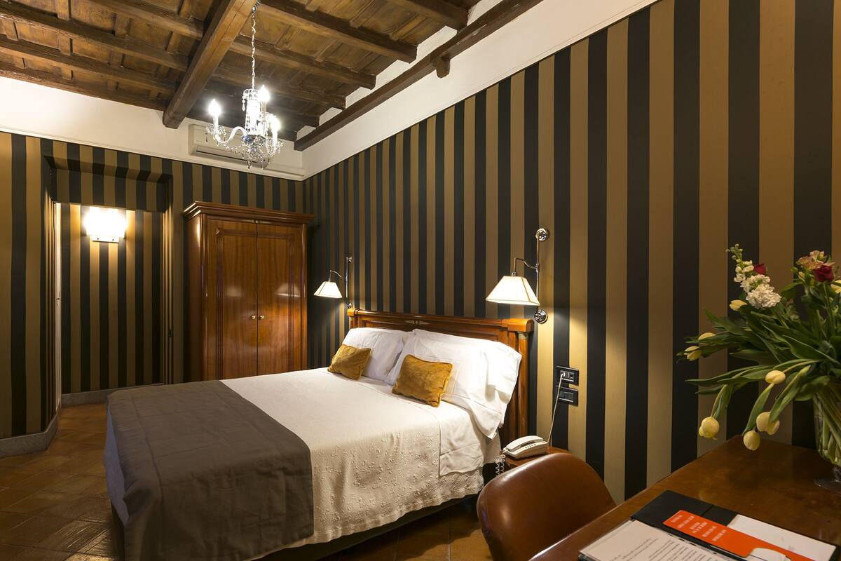 A hotel room with a bed, a wooden wardrobe, a desk with a chair, a telephone, and a vase with flowers.