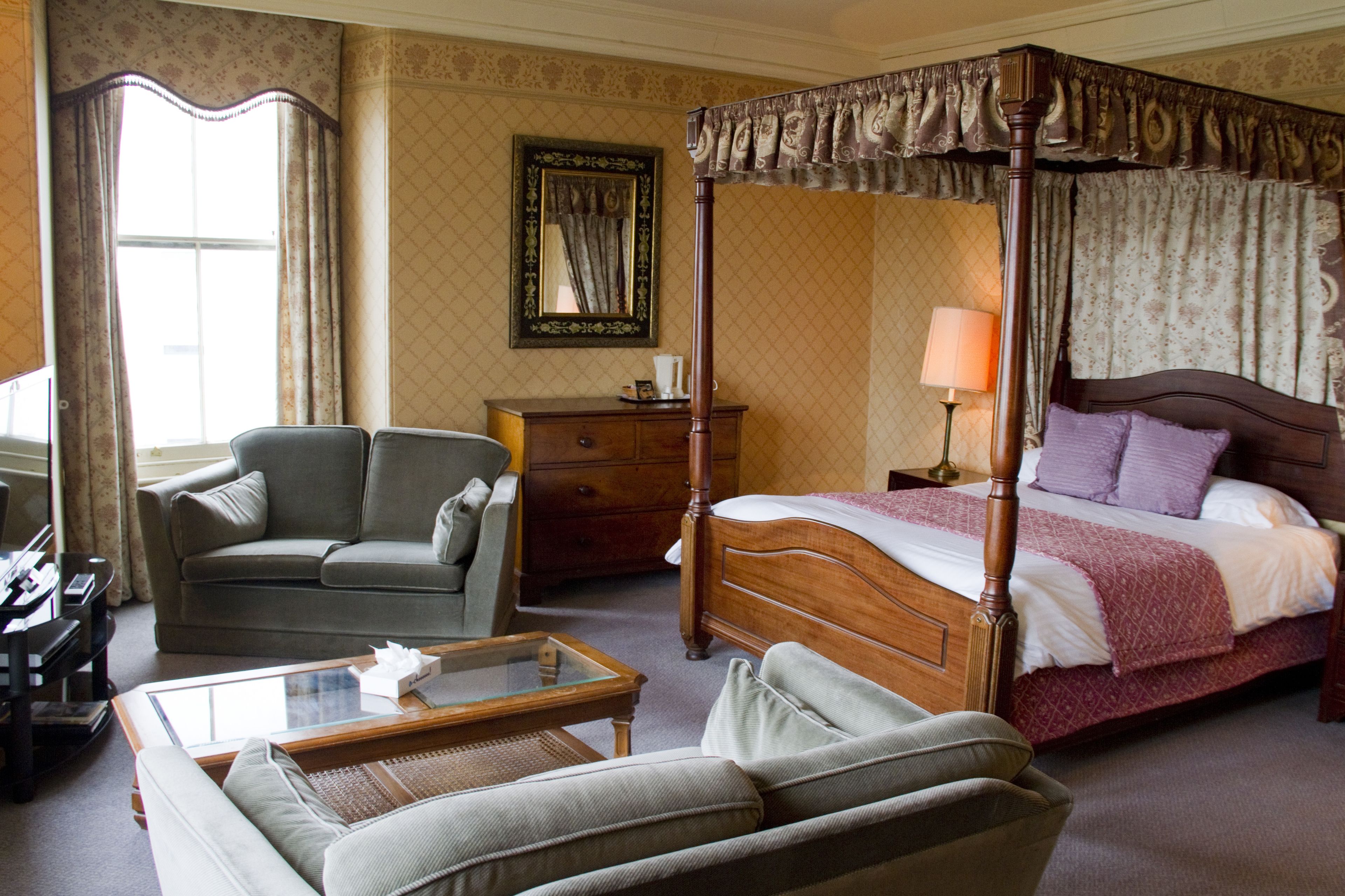 A four-poster canopy bed with a wooden frame and a red bedspread, a grey sofa, a wooden nightstand, and a glass coffee table in a room with patterned wallpaper and a large mirror on the wall.