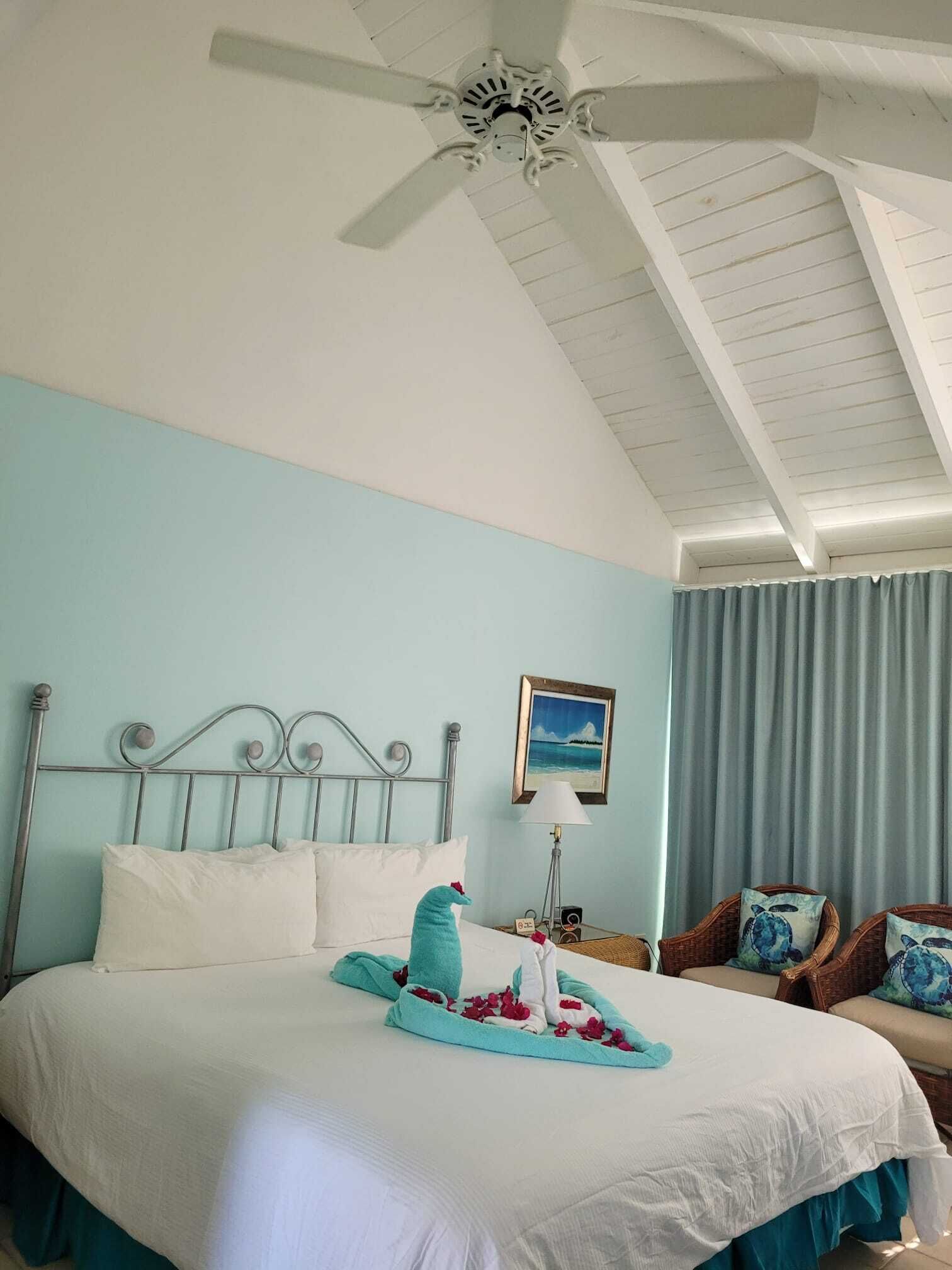 Habitación de hotel con cama, ventilador de techo, mesita de noche, lámpara y vista al océano a través de la ventana.