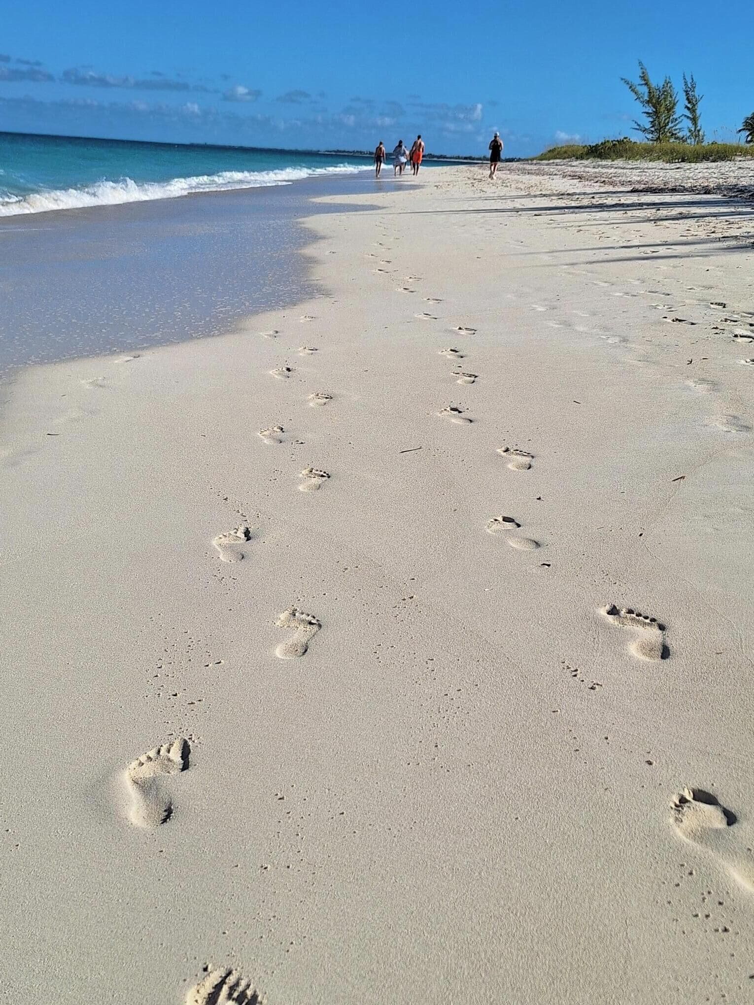 A sandy beach with footprints leading towards the ocean, a clear sky, and a few people in the distance.