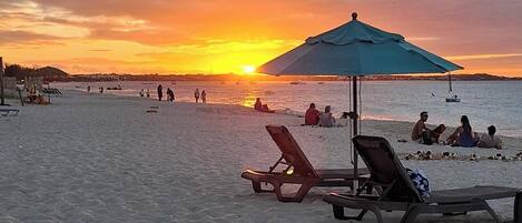 A beach with lounge chairs and umbrellas, people relaxing, and boats in the water.
