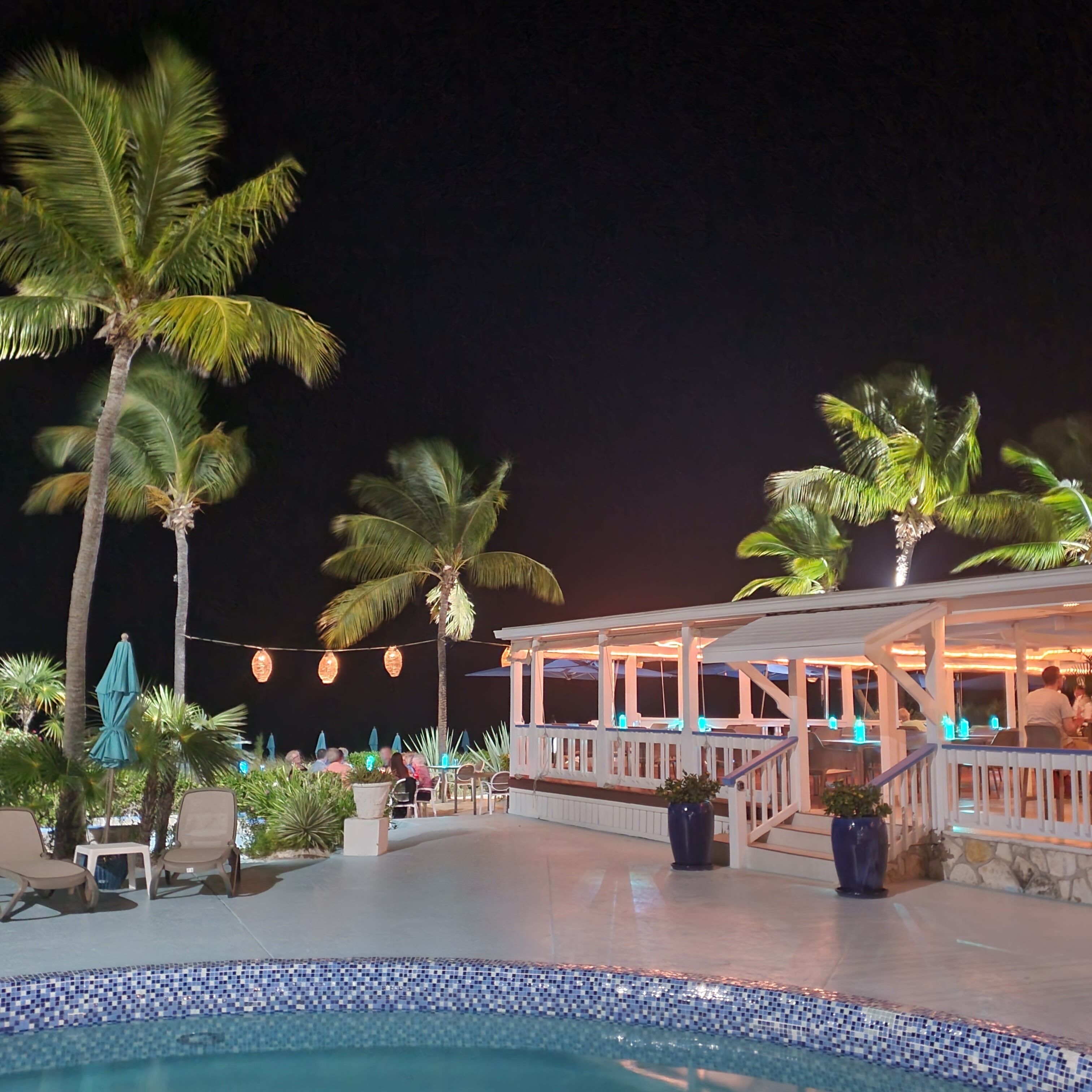 Área de piscina exterior de noche con palmeras, una zona de estar cubierta y un borde de piscina con baldosas.