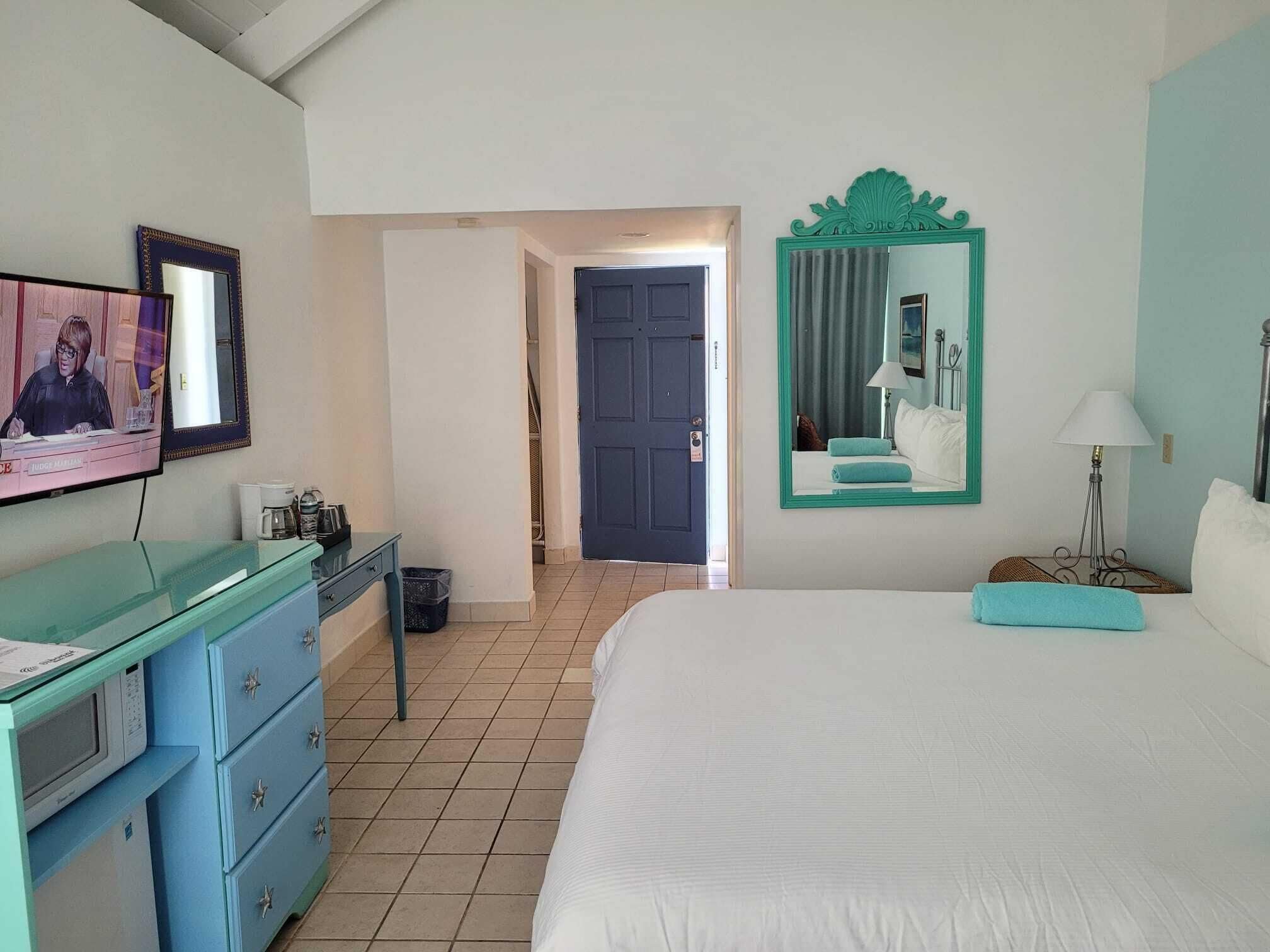 A hotel room with a bed, a desk with a computer, a mirror, and a door leading to another room.