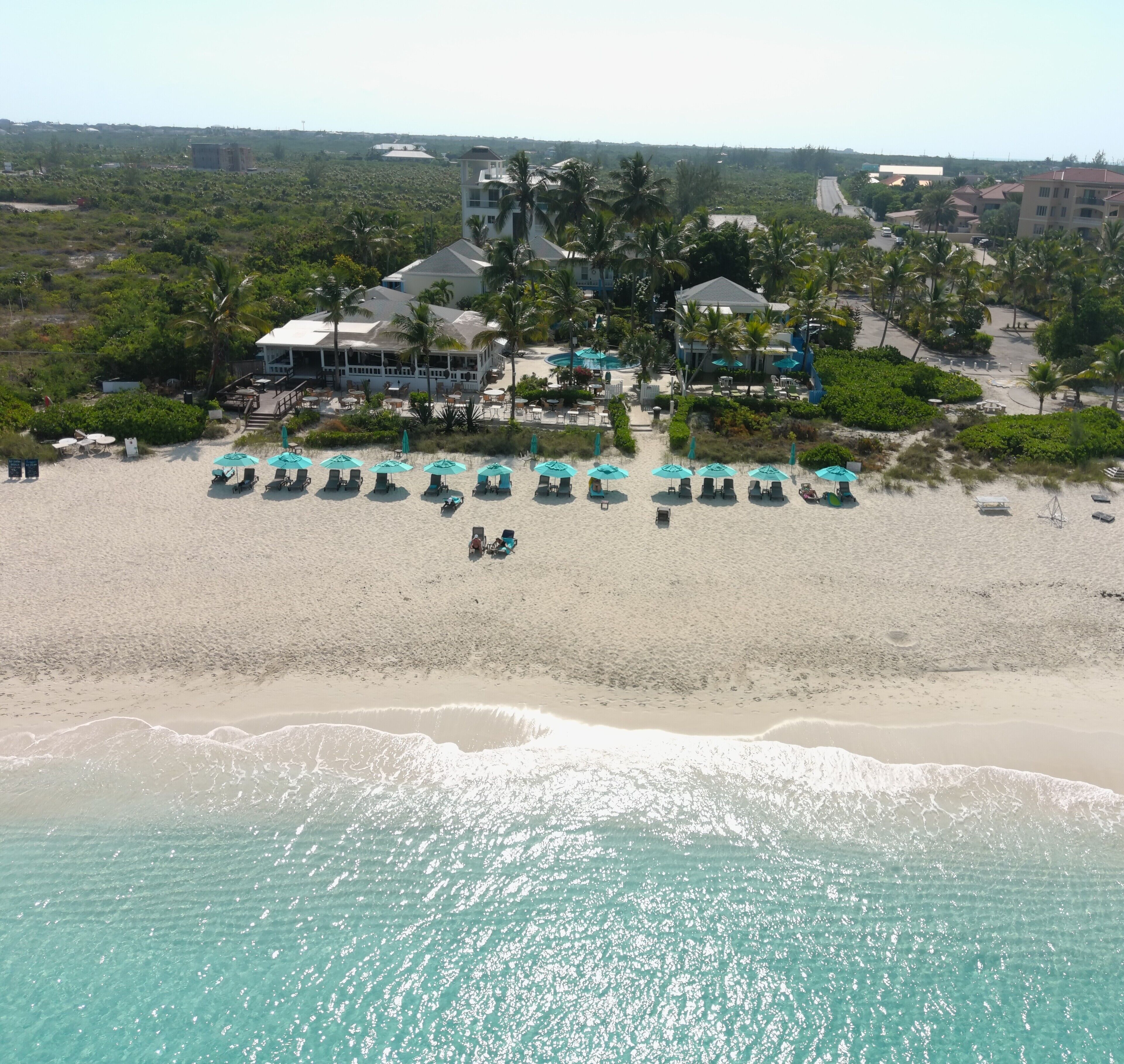 A beach resort with multiple lounge chairs and umbrellas, a pool area, and a building with a covered patio.