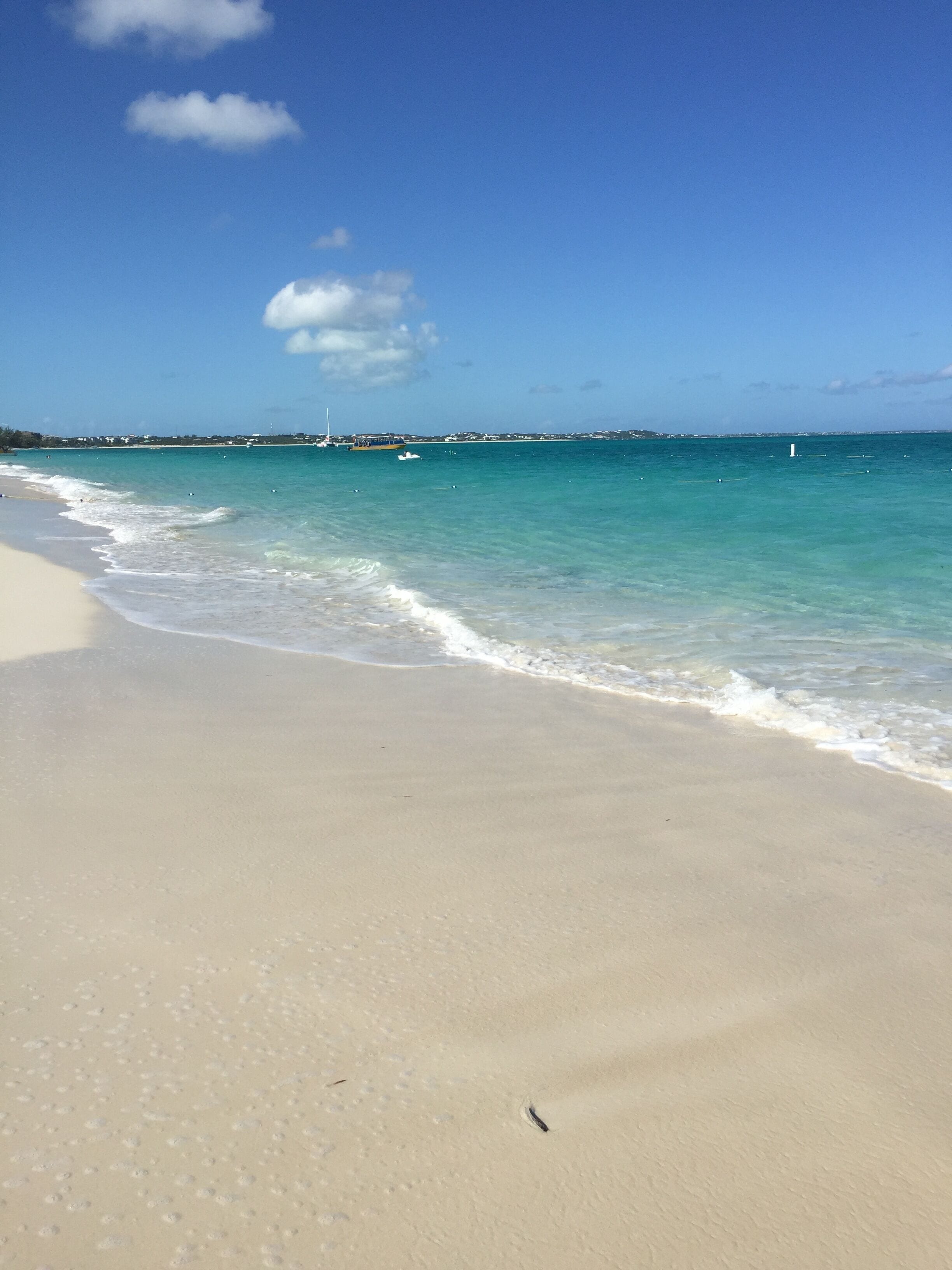 A beach with clear turquoise water, a sandy shore, and a distant shoreline with buildings and boats.