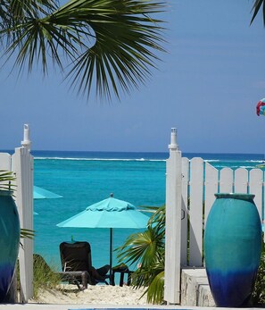 A beach view with a white fence, two large vases, a blue umbrella, and a chair. - Sibonné Beach Hotel (Providenciales)