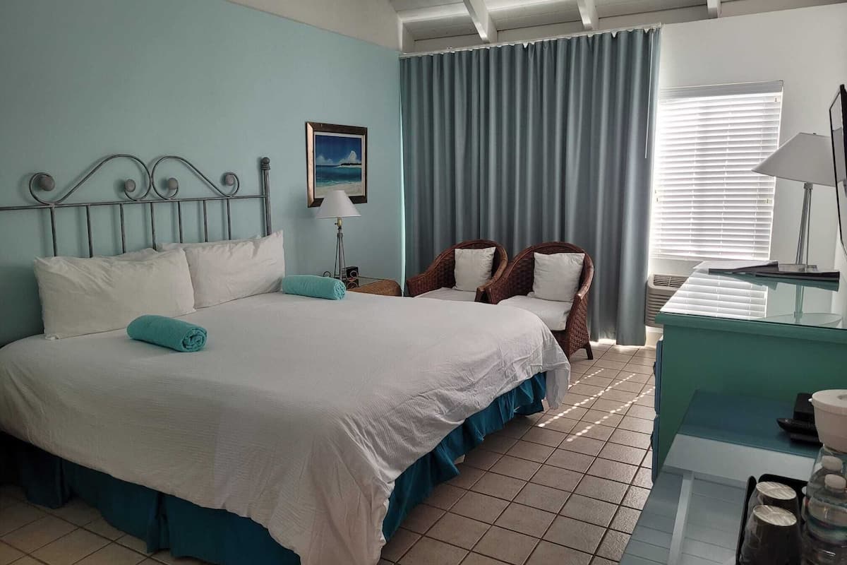 A bedroom with a large bed, two wicker chairs, a desk with a lamp, and a window with blinds.