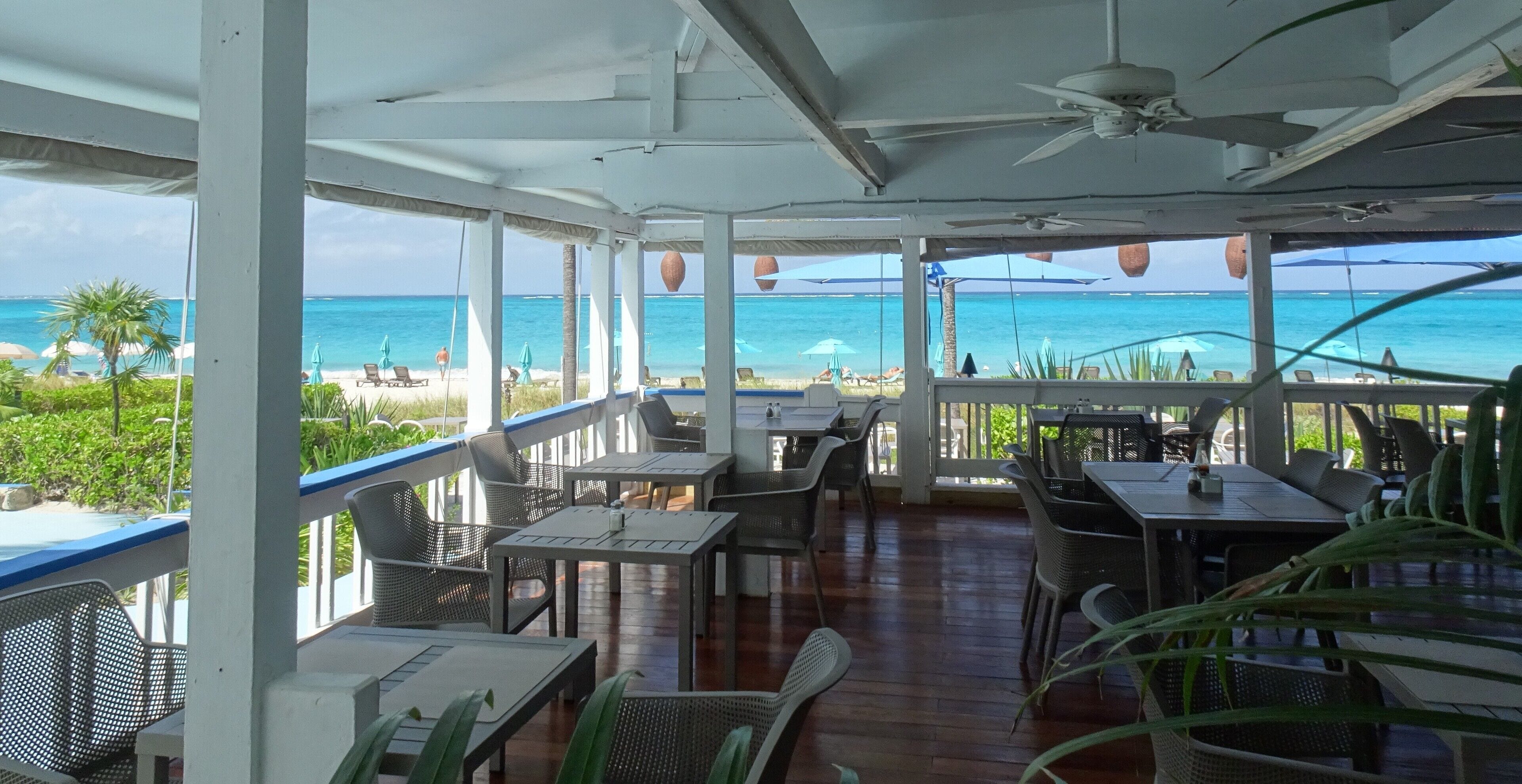 A covered outdoor seating area with tables and chairs, overlooking a beach with clear blue water.
