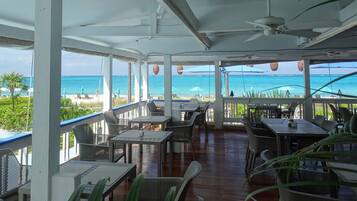 A covered outdoor seating area with tables and chairs, overlooking a beach with clear blue water.