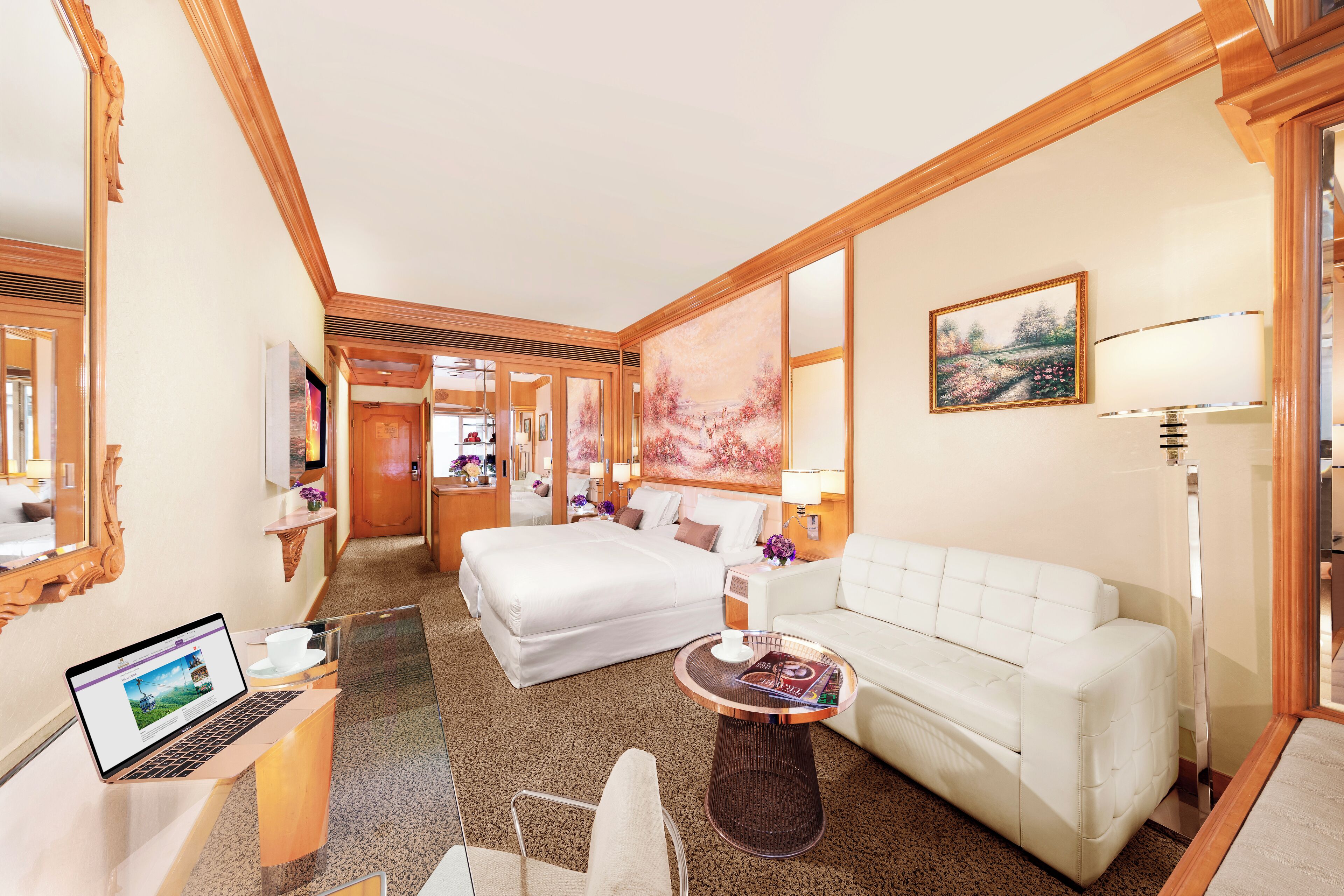 Une chambre d’hôtel avec un lit, un canapé blanc, un bureau avec un ordinateur portable, une table basse et un tableau au mur.