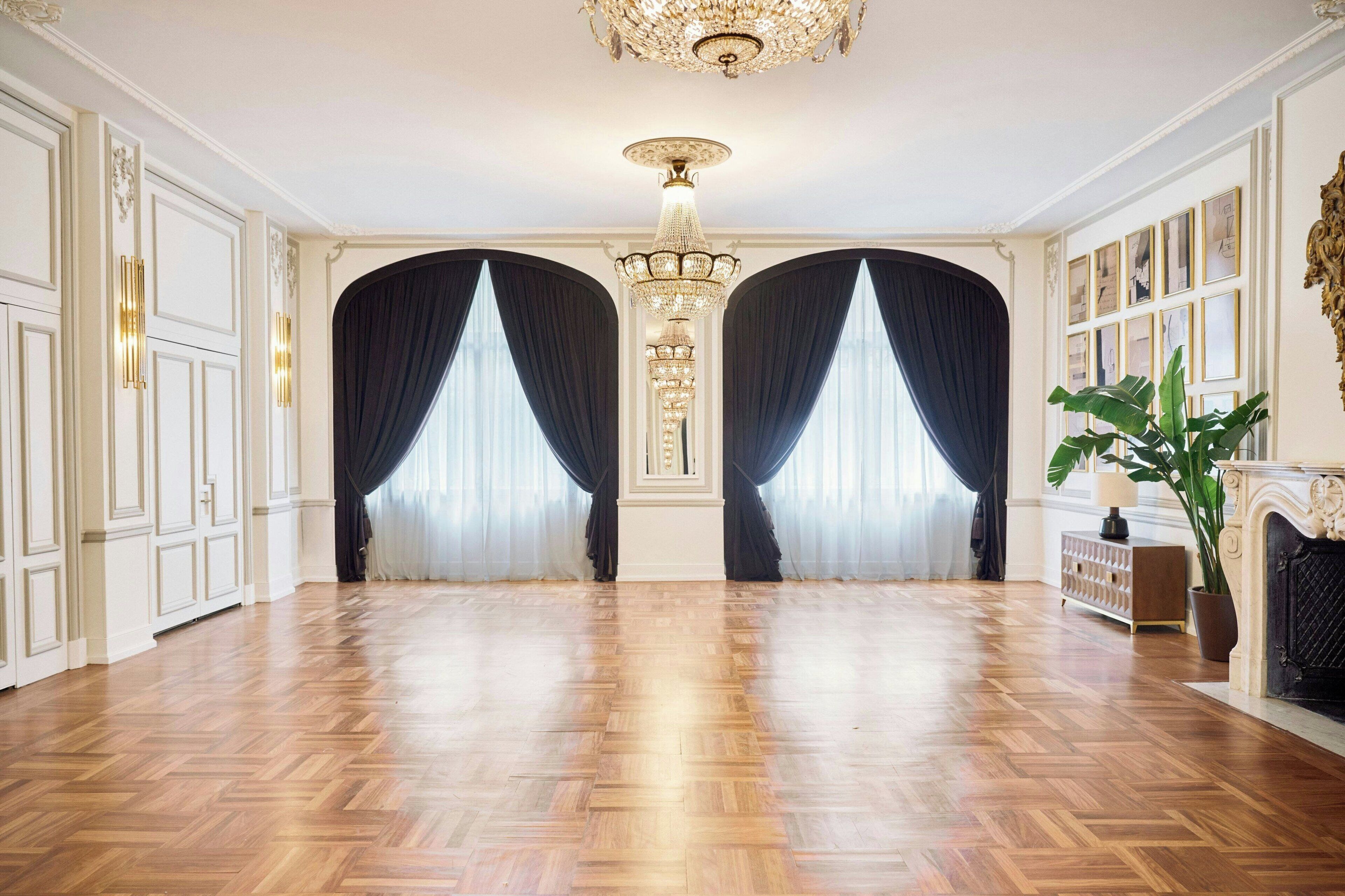 A spacious room with a polished wooden floor, a grand chandelier, and dark curtains framing a large window.