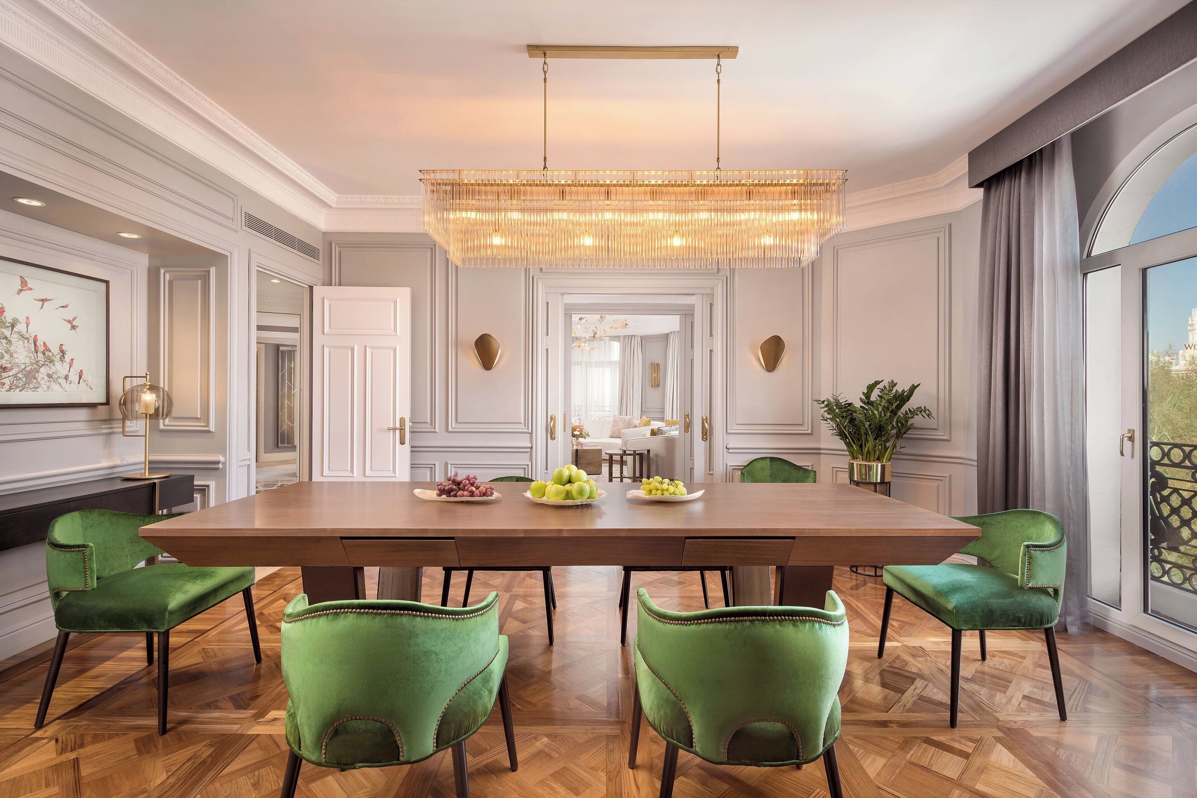 A dining room with a large wooden table, green upholstered chairs, a chandelier, and a view of a building through a large window.
