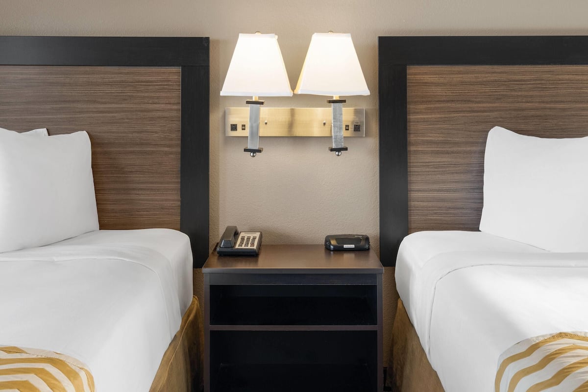 A hotel room with two beds, a nightstand with a lamp, a phone, and a remote control.